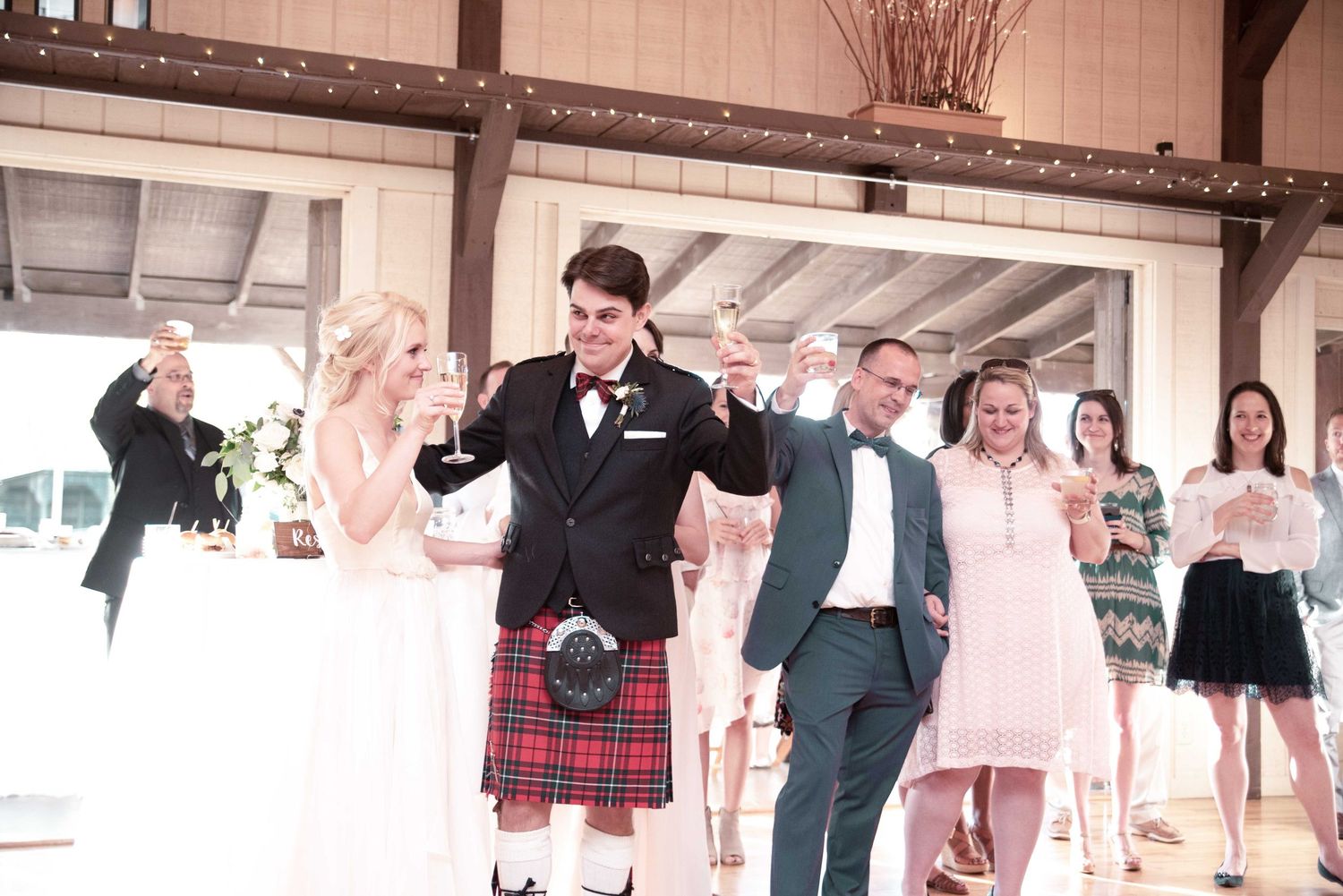 Joyful wedding reception with groom in red tartan kilt and bride in white gown celebrating with guests in elegant venue.