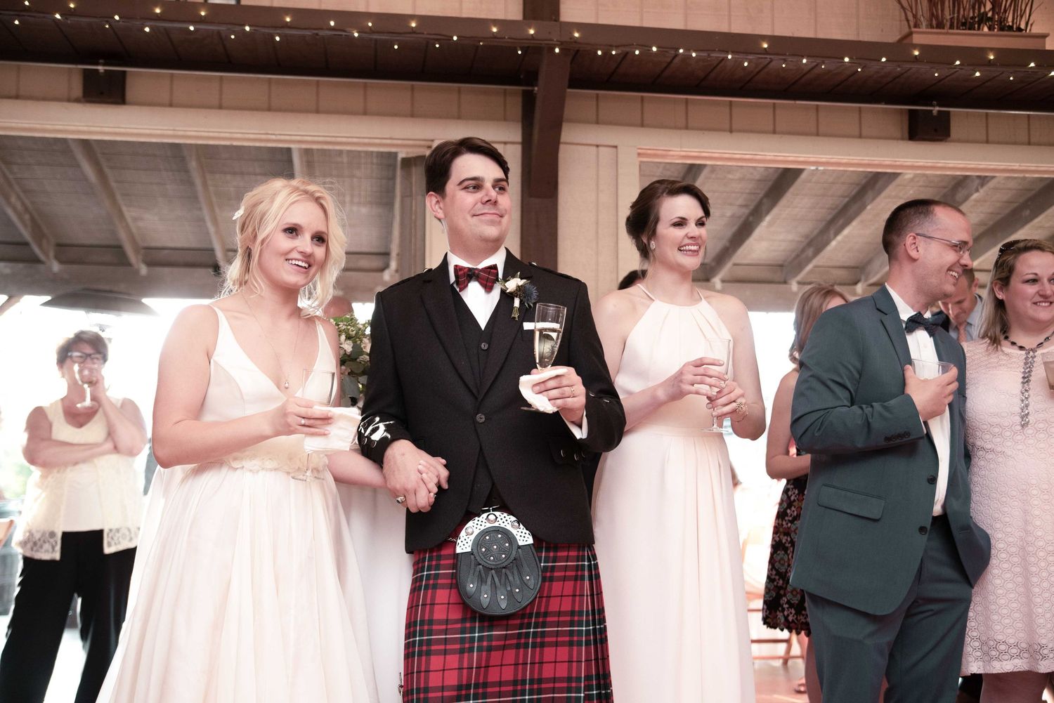 Wedding party in Scottish attire, groom in kilt raising a toast with bridesmaids in white gowns at rustic venue.