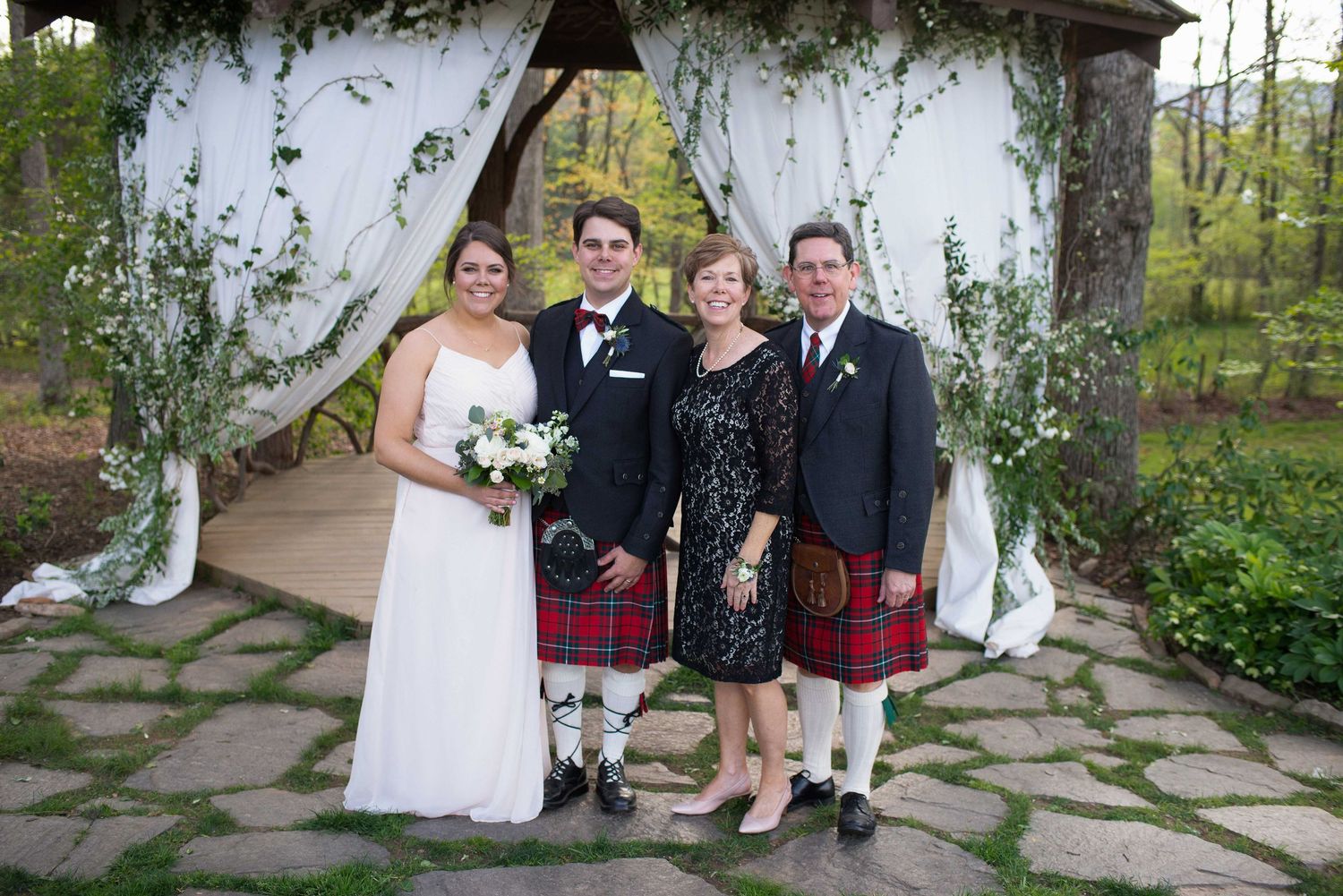 Bride and groom with parents in Scottish kilts celebrate at elegant outdoor garden wedding.