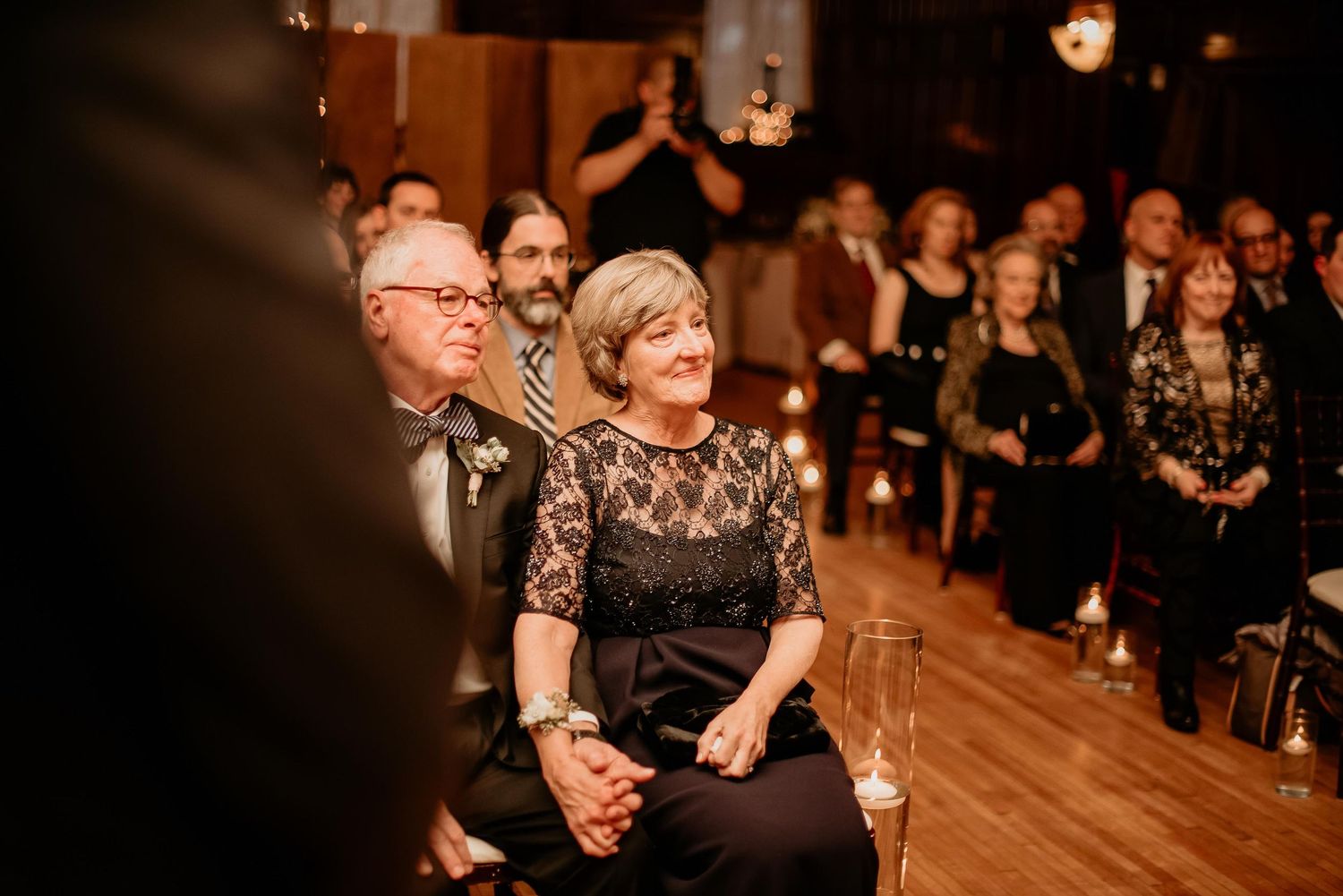 Guests smile warmly during a wedding reception in a warmly lit venue, dressed in elegant formal attire.