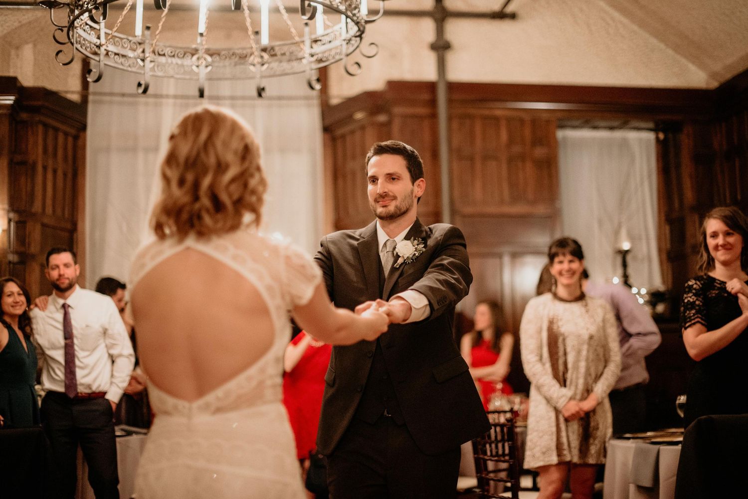 Groom smiles at bride during first dance at rustic indoor wedding reception with guests watching.