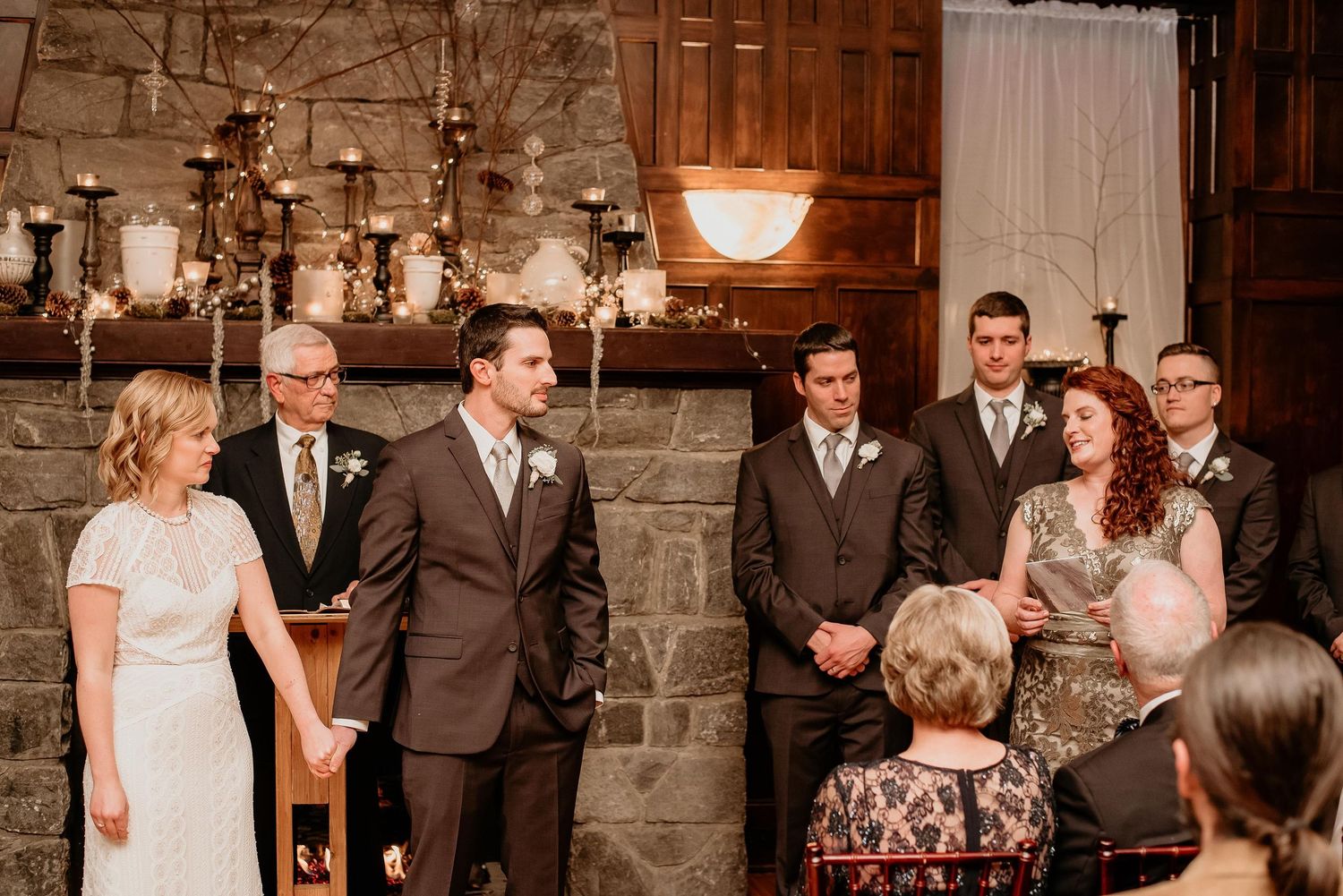 Wedding ceremony in a stone venue with groom, groomsmen, and guests gathered near a candlelit fireplace.