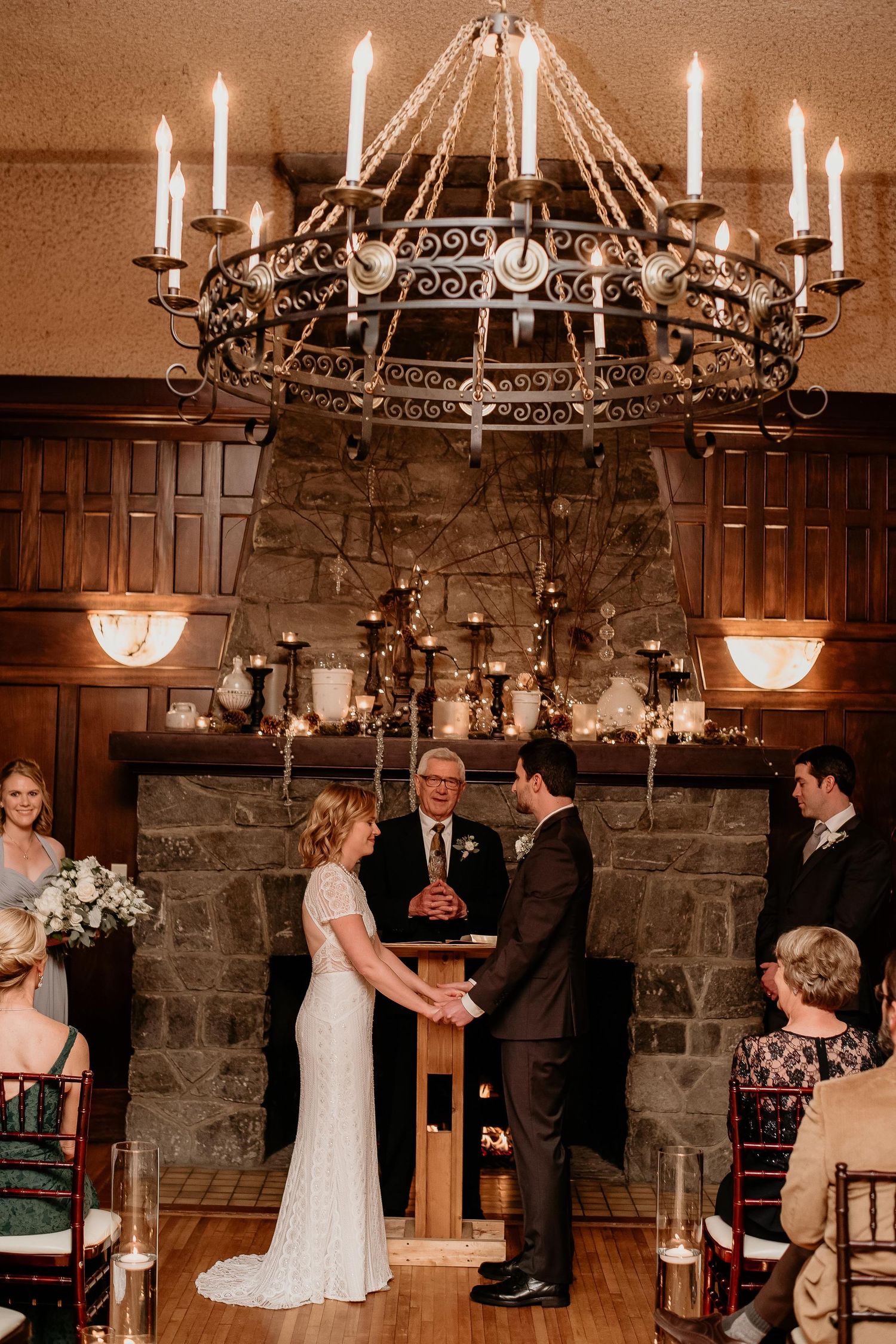 Couple exchanging vows at elegant indoor wedding ceremony with stone fireplace and ornate chandelier.
