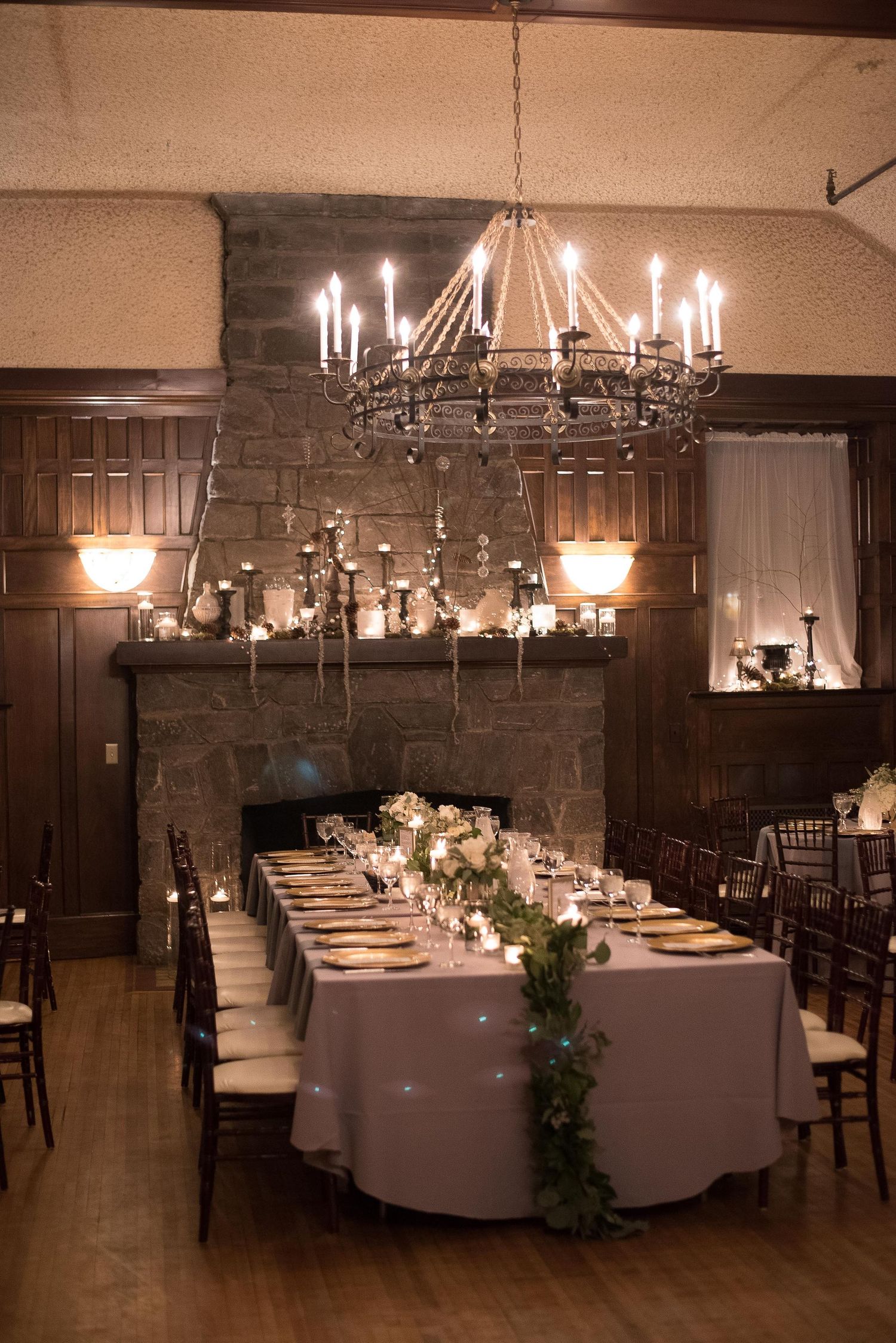 Elegant wedding reception with long dining table, greenery garland, candle chandelier, and stone fireplace in a warmly lit venue.