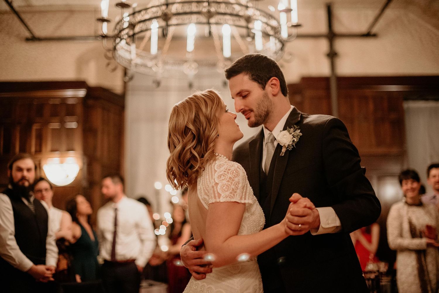 Bride and groom share romantic first dance under chandelier at wedding reception with guests watching.