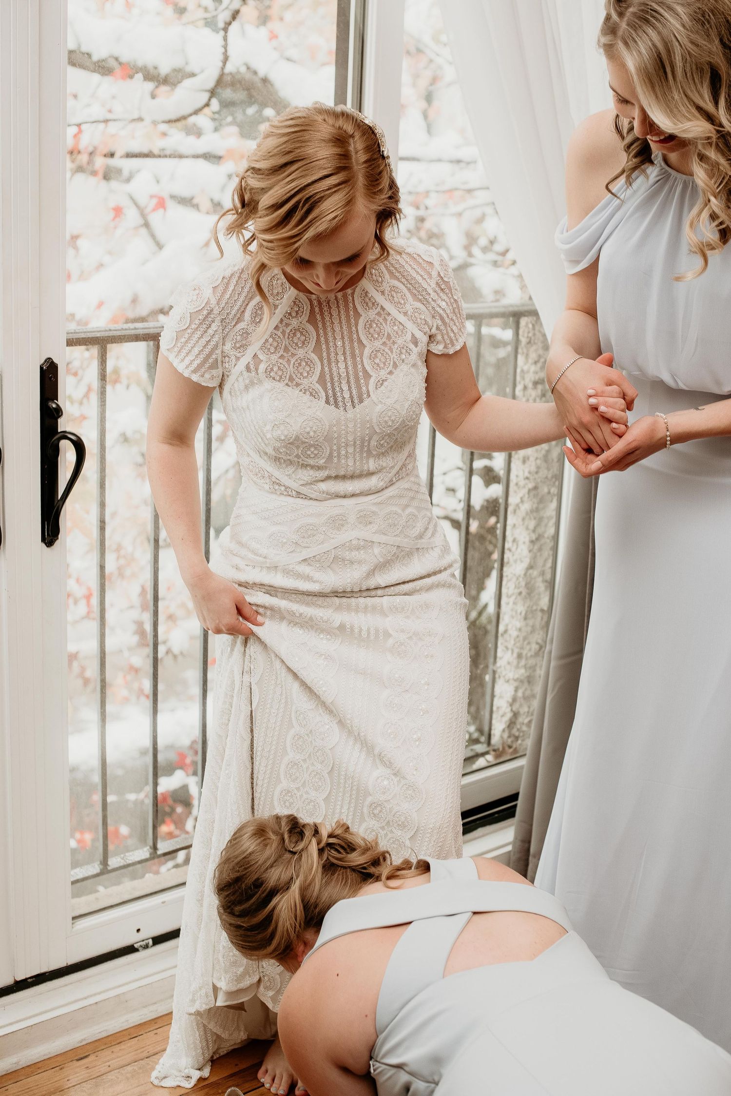 Bride in lace wedding dress being helped by bridesmaids in grey dresses near a sunlit window.