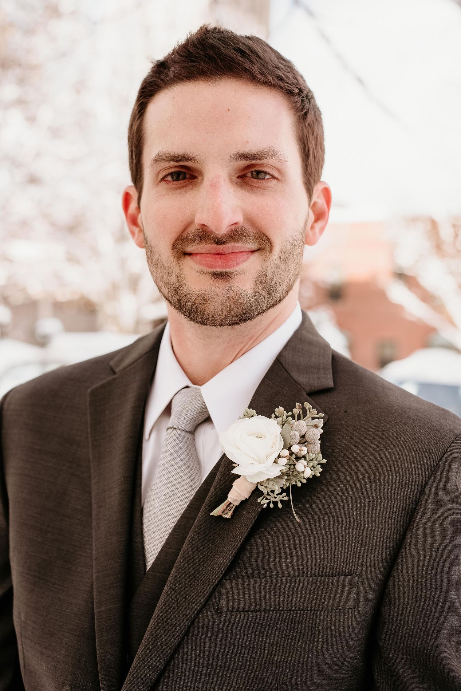 A smiling groom in a gray suit with a white rose boutonniere poses for a formal wedding portrait outdoors.