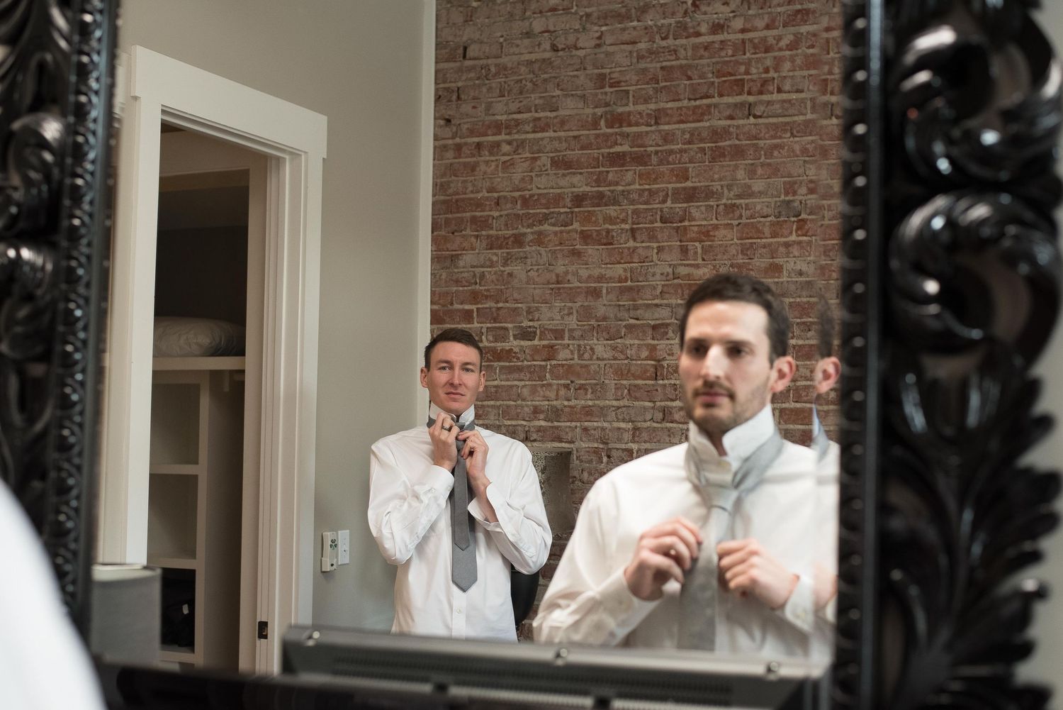 Two groomsmen in white dress shirts adjust their ties while getting ready, reflected in an ornate black mirror.