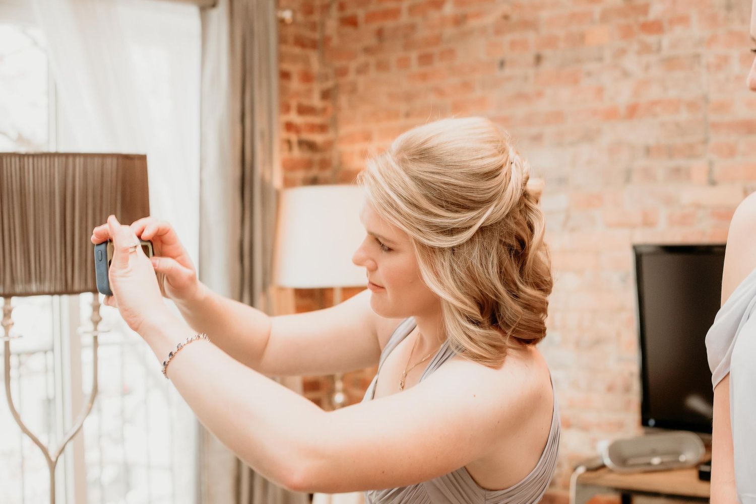Blonde woman with elegant updo hairstyle adjusting a lamp shade in a rustic brick-walled room.