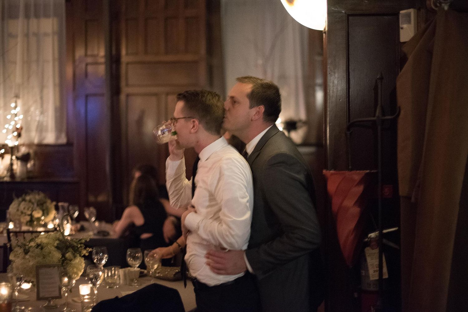 Two men in formal attire stand together at an elegant indoor event with candlelit tables and decorative lighting.