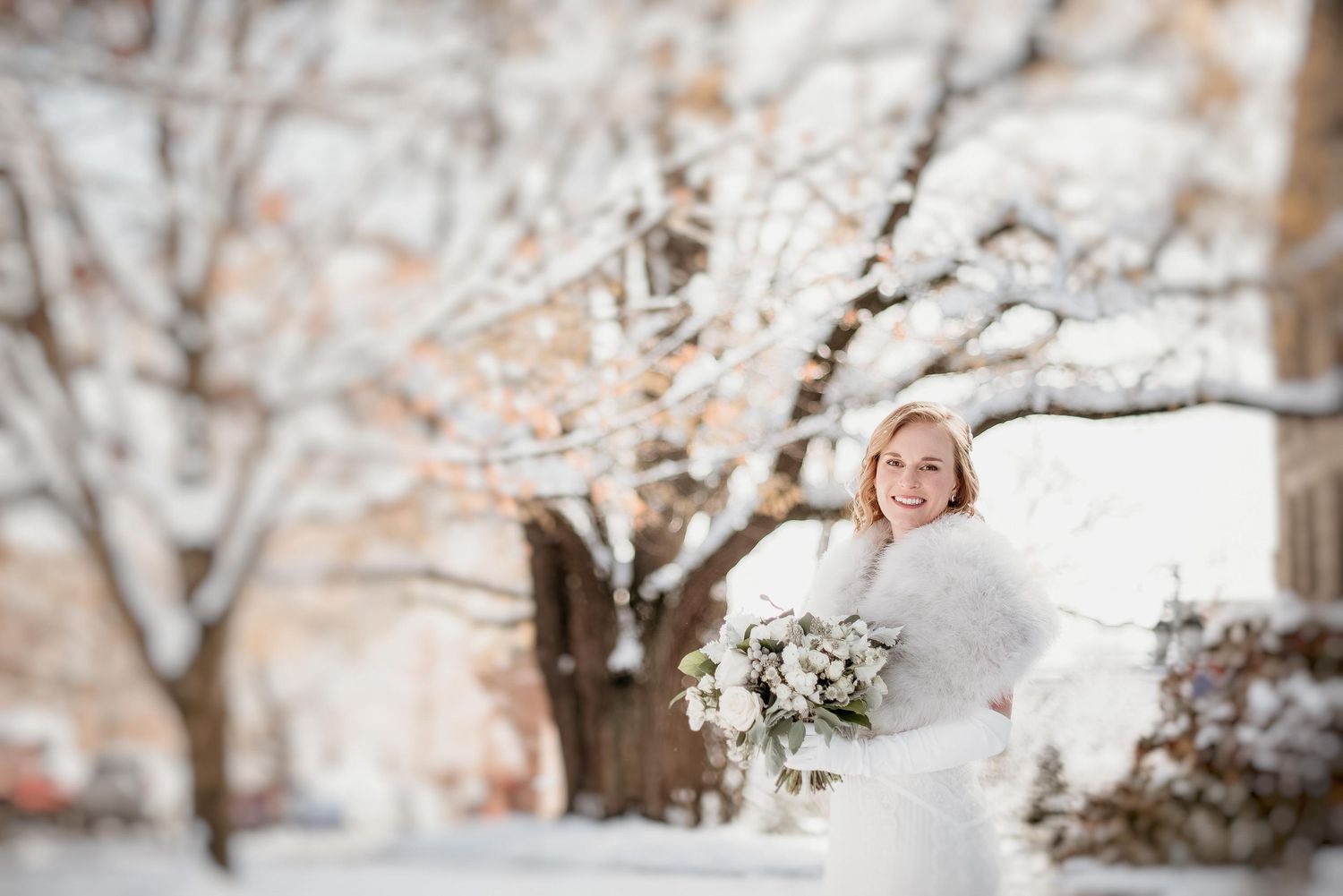 Smiling bride in white dress and fur wrap holding bouquet in snowy winter outdoor setting.