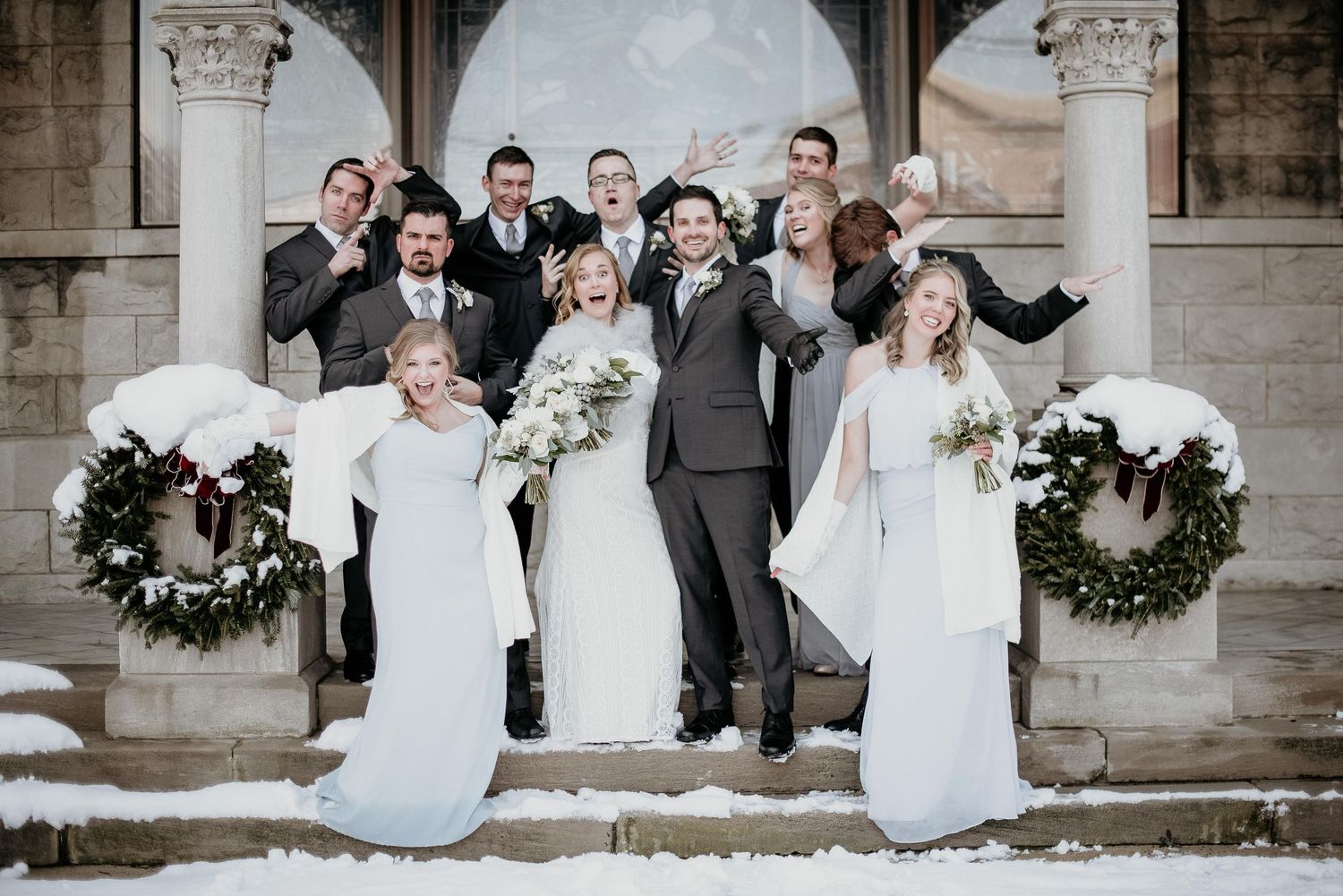 Wedding party celebrating on snowy church steps in winter, bridesmaids in white gowns and groomsmen in gray suits.