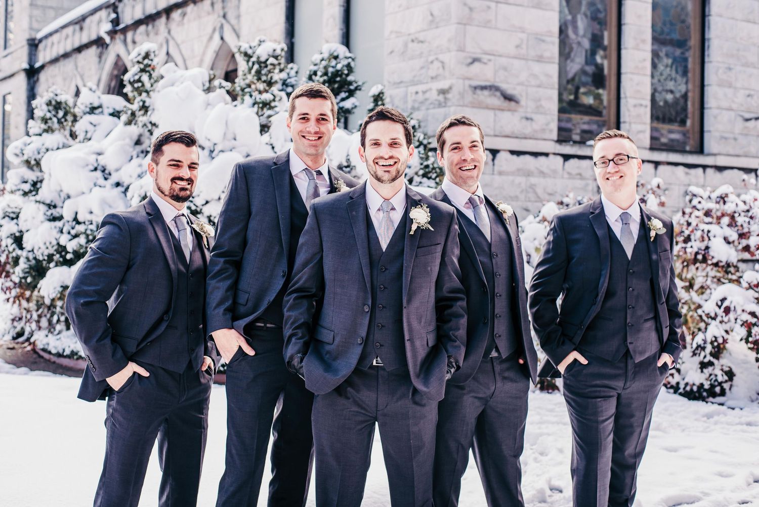 Five groomsmen in navy suits pose outdoors in winter snow near a stone building with snow-covered evergreen trees.