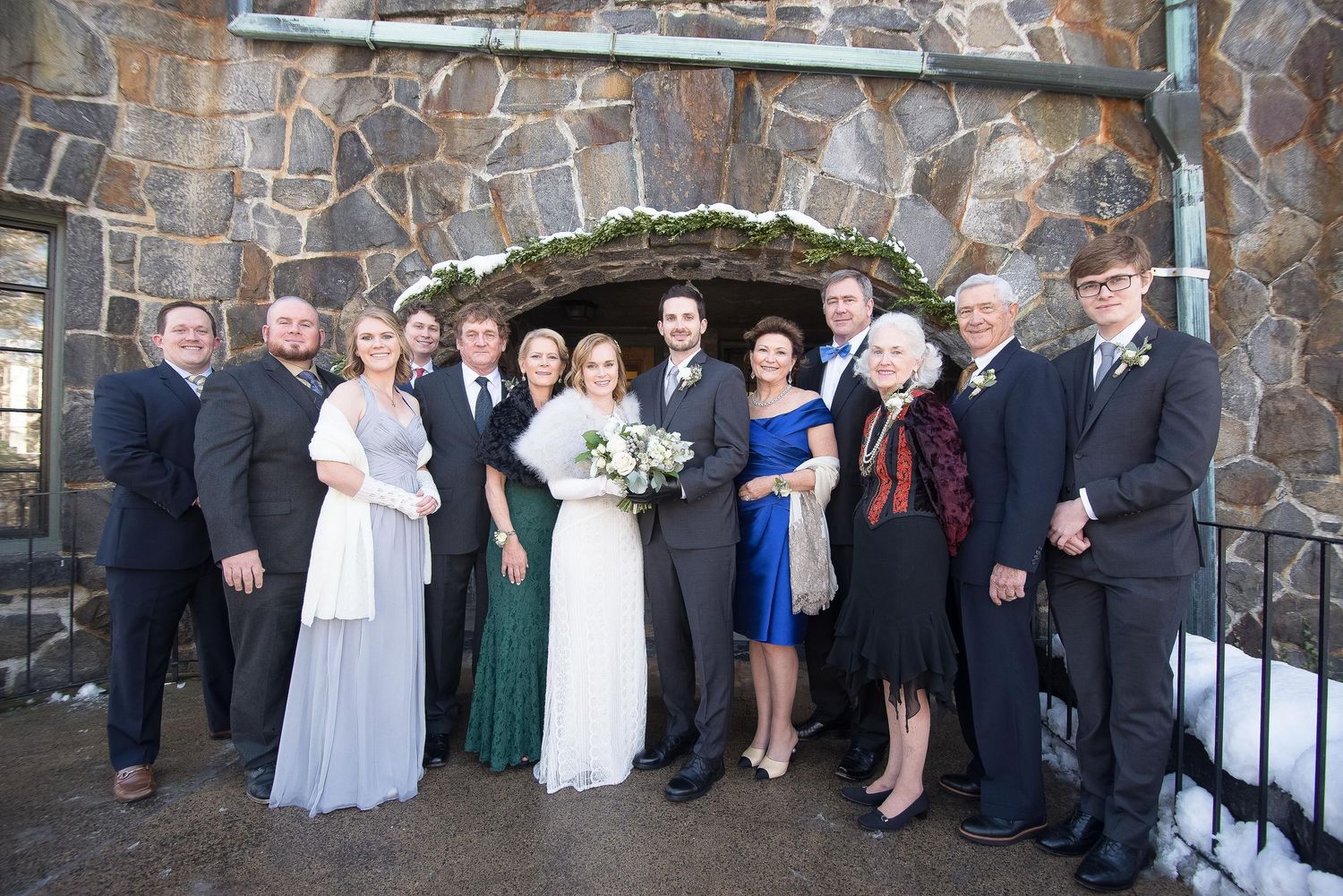Wedding party poses in formal attire before a stone arch backdrop in a winter outdoor setting.