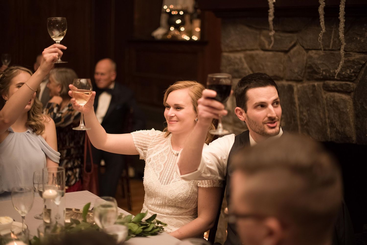 Wedding guests raising glasses in a toast at an elegant reception near a stone fireplace.