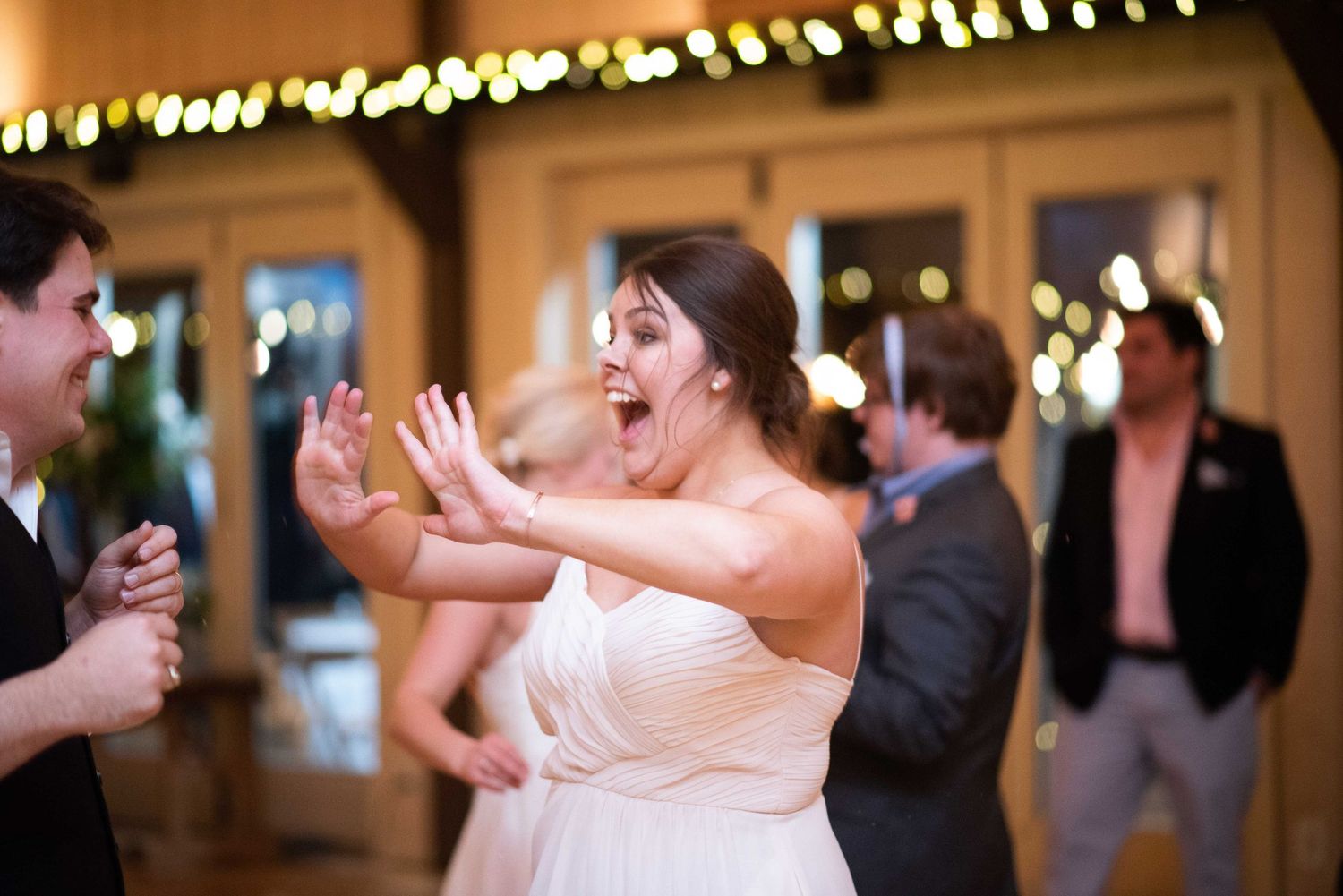 Bride in white gown dancing joyfully at wedding reception with bokeh lights in background.