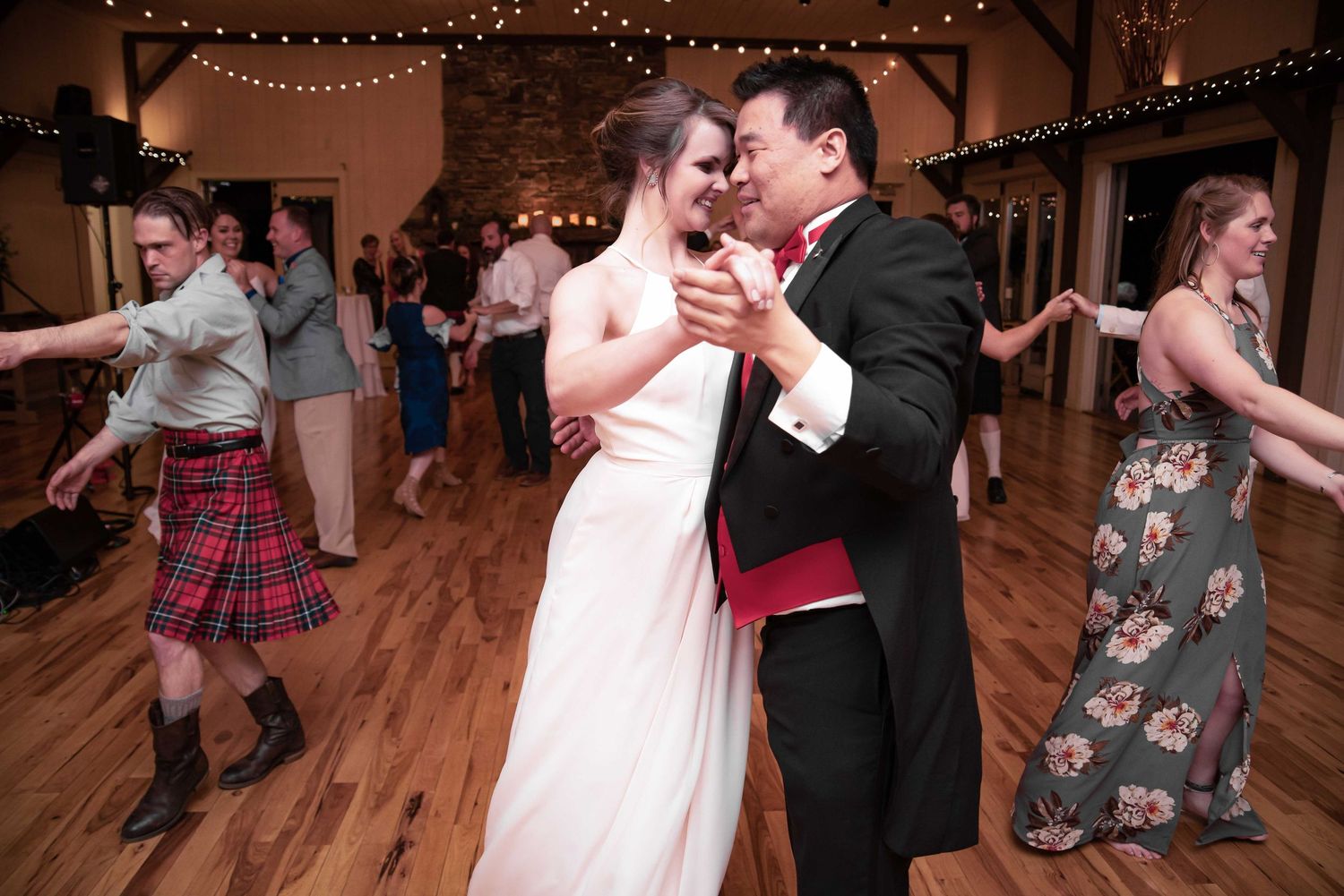 Bride and groom share a romantic first dance at rustic wedding reception with guests dancing in background.