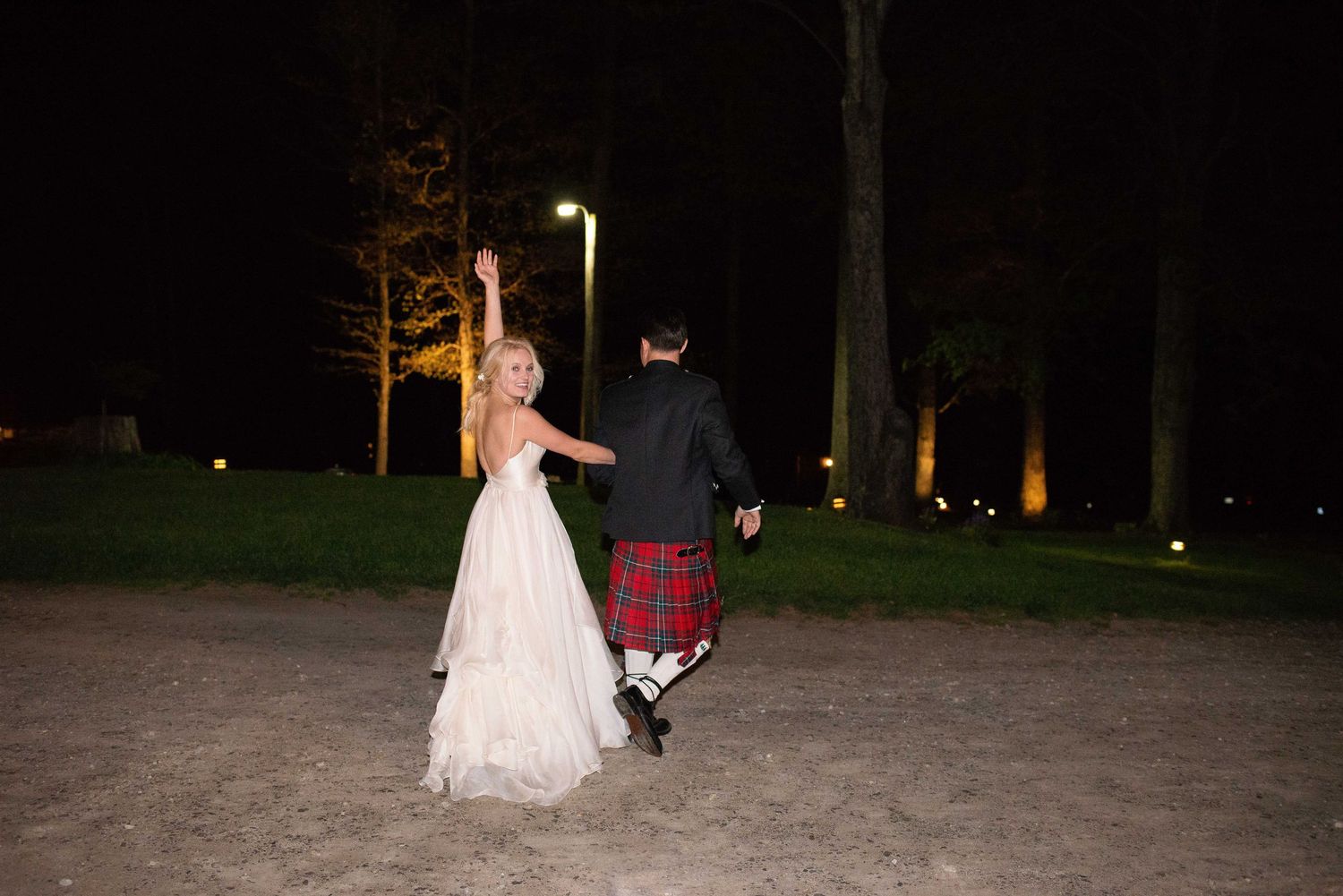 Bride in white gown and groom in red tartan kilt celebrate outdoors at night, raising arms joyfully.