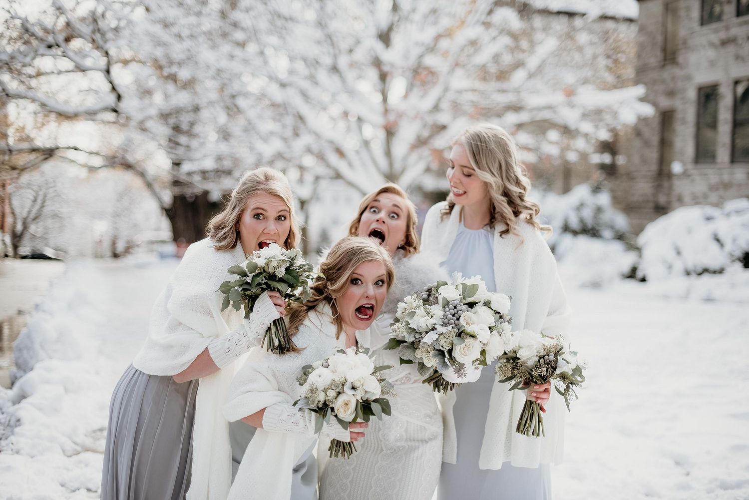 Four bridesmaids in white wraps holding bouquets laugh and pose playfully in a snowy winter wedding outdoor setting.