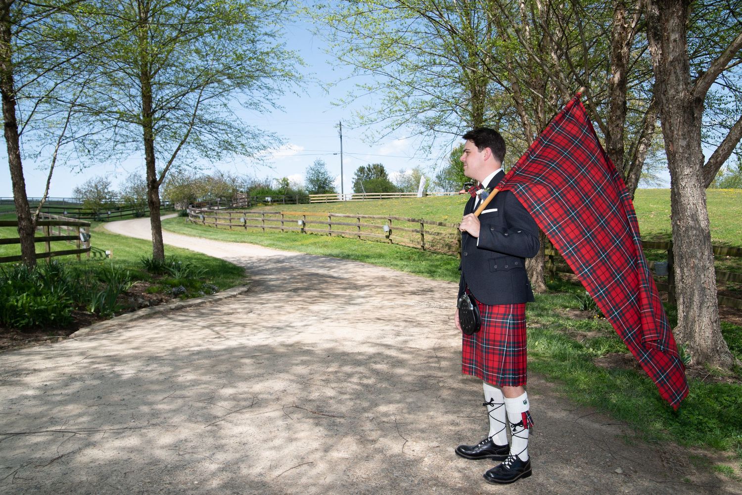 Scottish bagpiper in traditional red tartan kilt standing on a scenic rural pathway surrounded by spring trees.