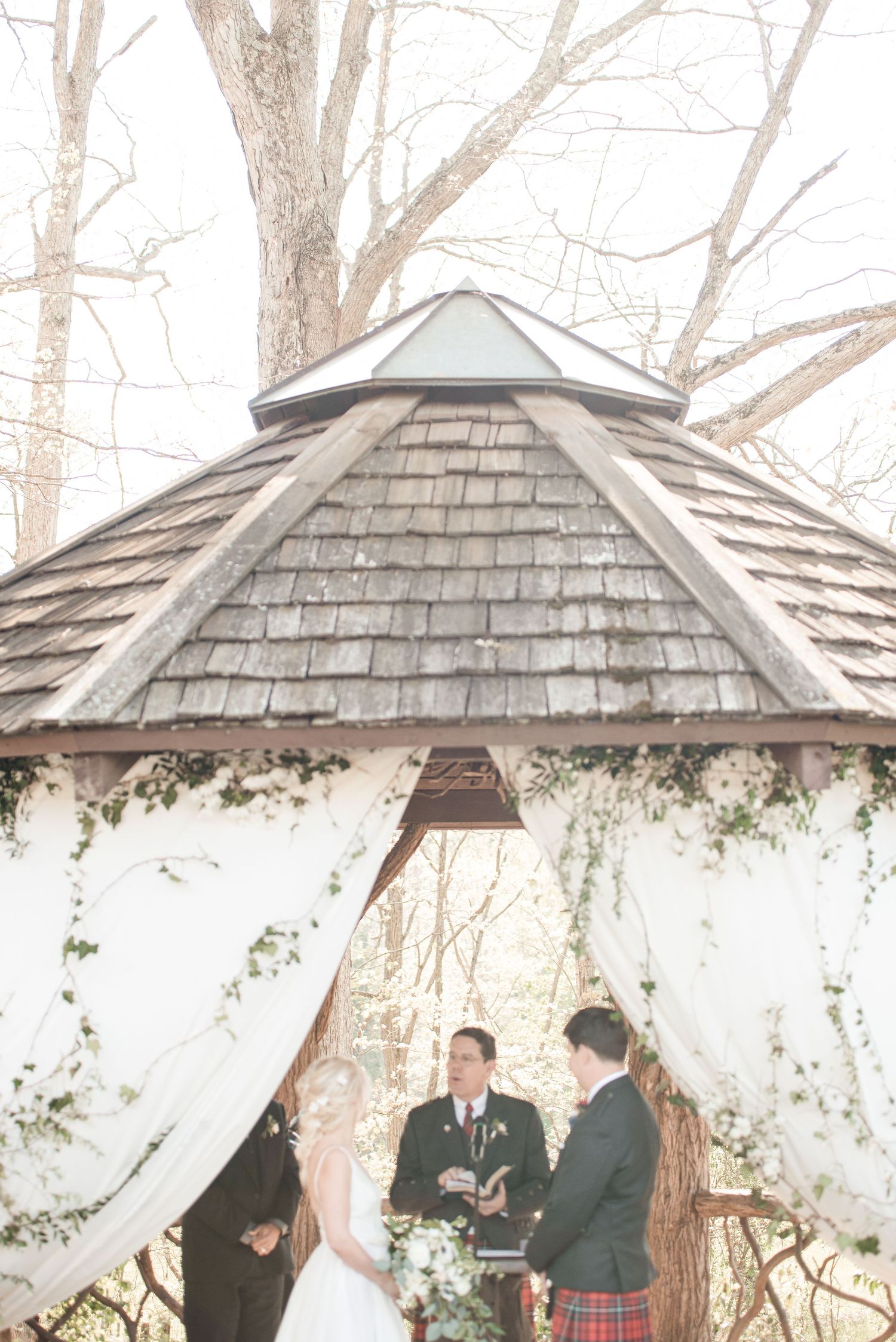 Couple exchanging vows under rustic gazebo adorned with white drapes and greenery.