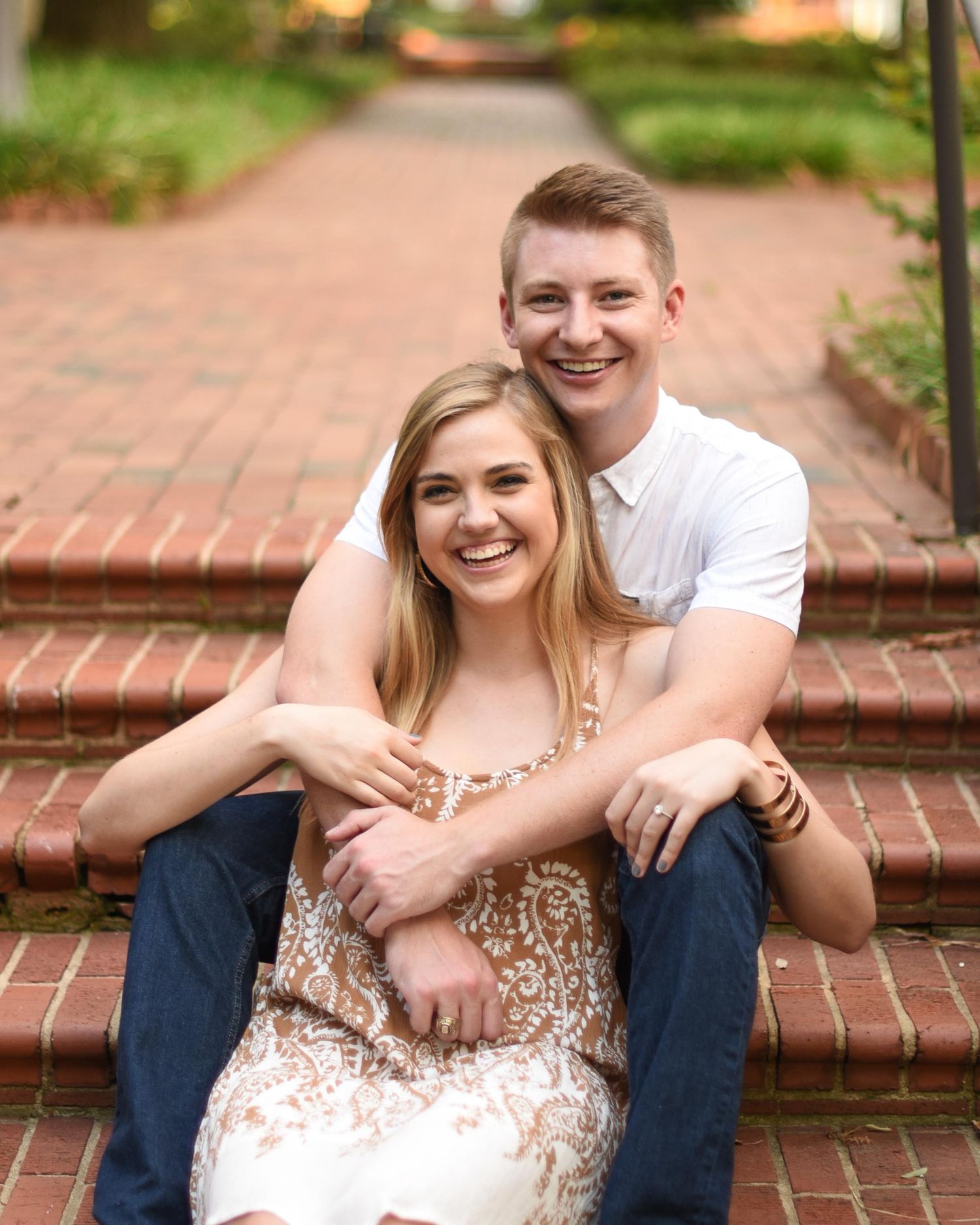 Smiling couple sitting on brick steps, man embracing woman from behind in an outdoor setting.