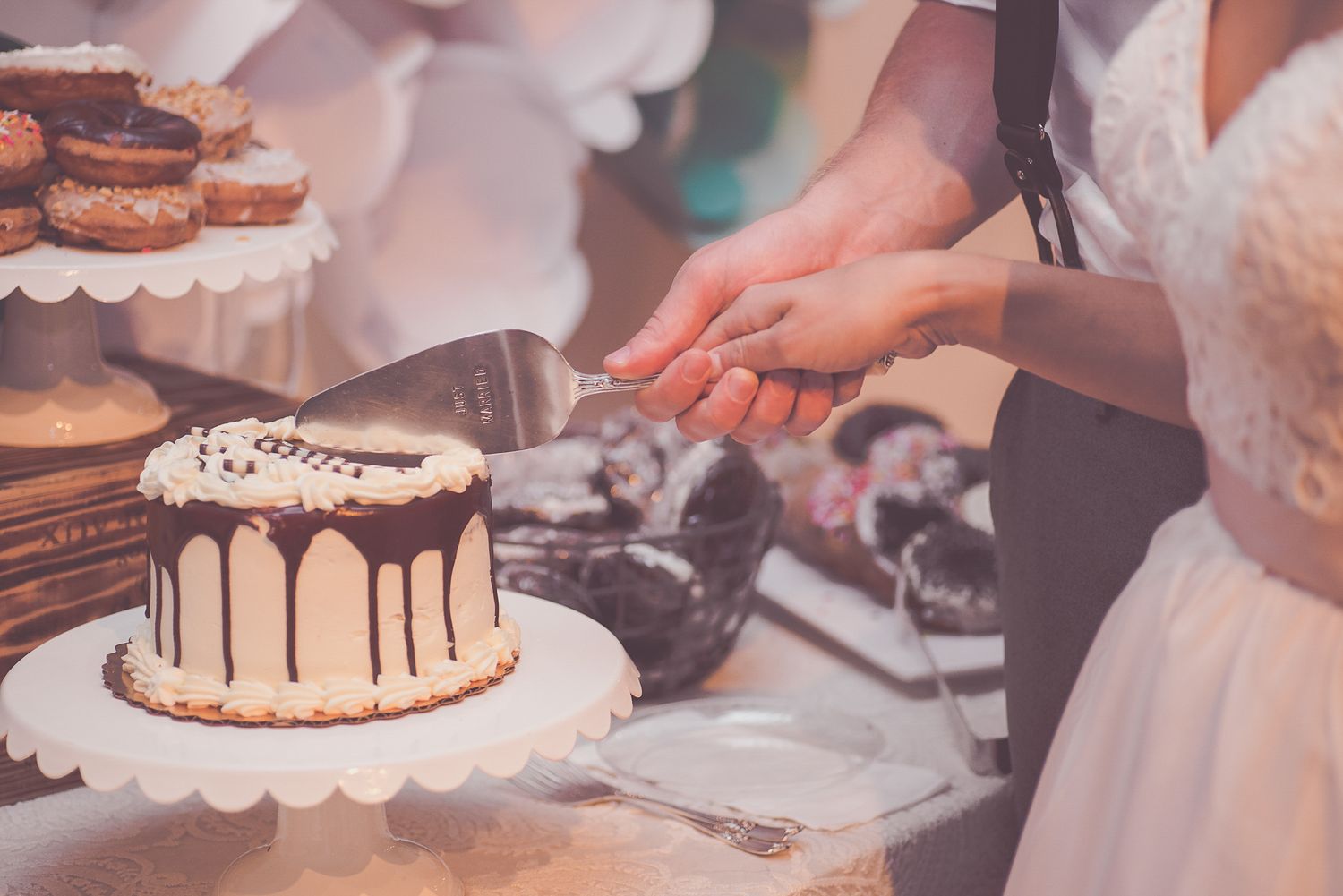Bride and groom cutting a chocolate drip wedding cake at a dessert table with donuts and treats.