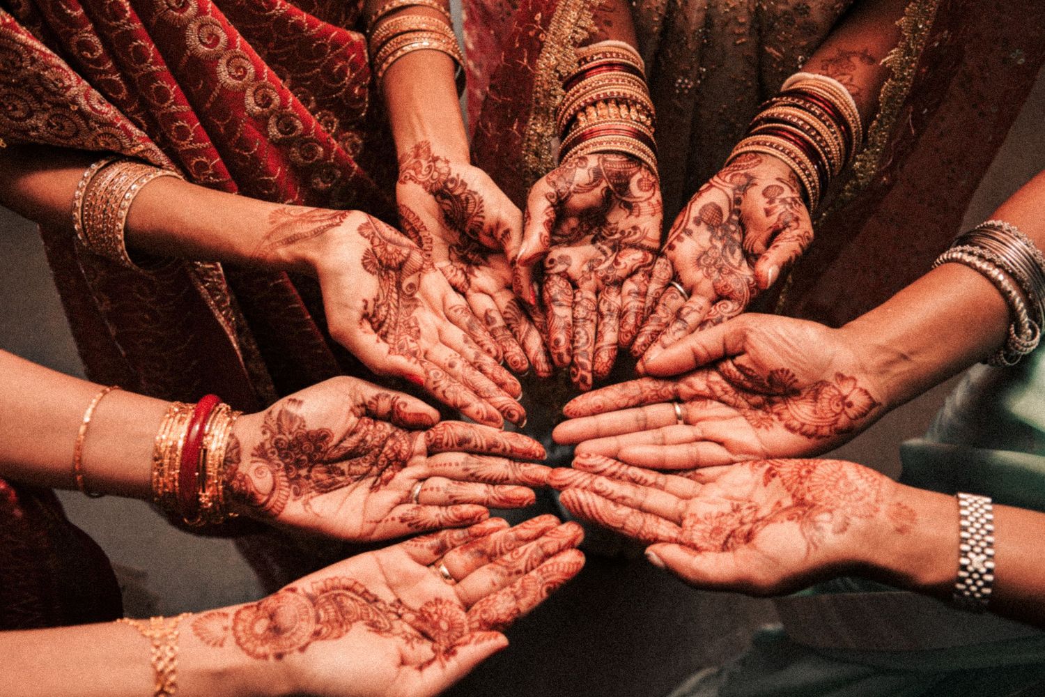 Multiple hands adorned with intricate henna mehndi designs and gold bangles at an Indian wedding.