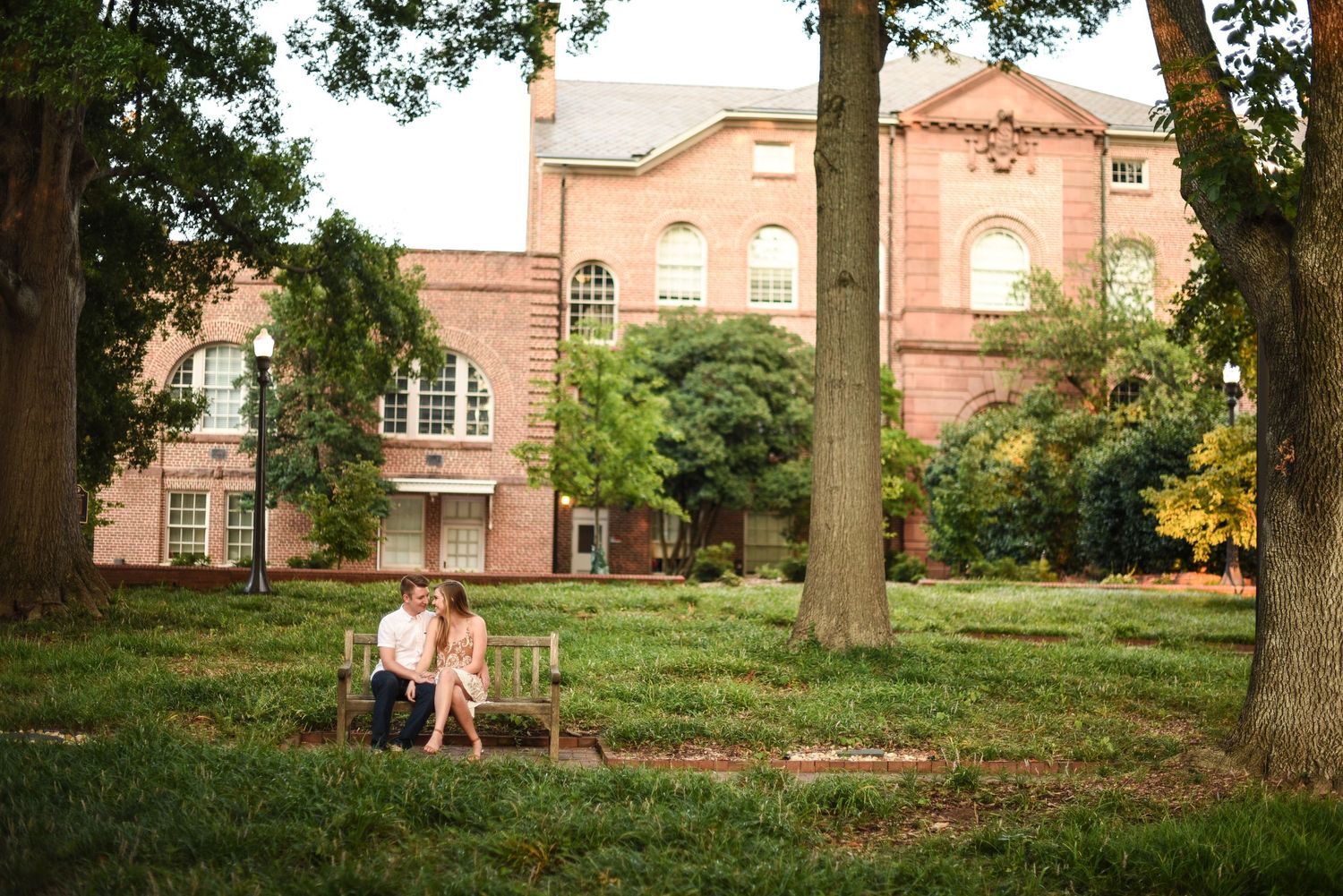 Couple sitting on a bench in a lush green park with a historic brick building backdrop.