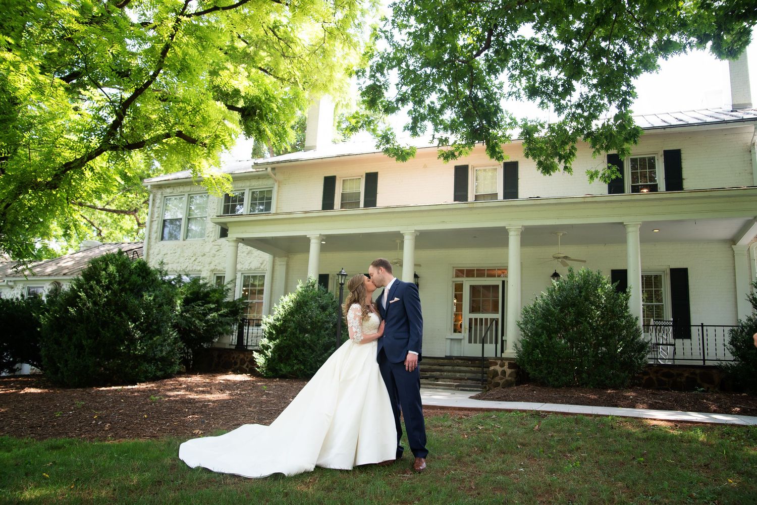 Bride and groom sharing a romantic kiss in front of a white colonial wedding venue surrounded by trees.