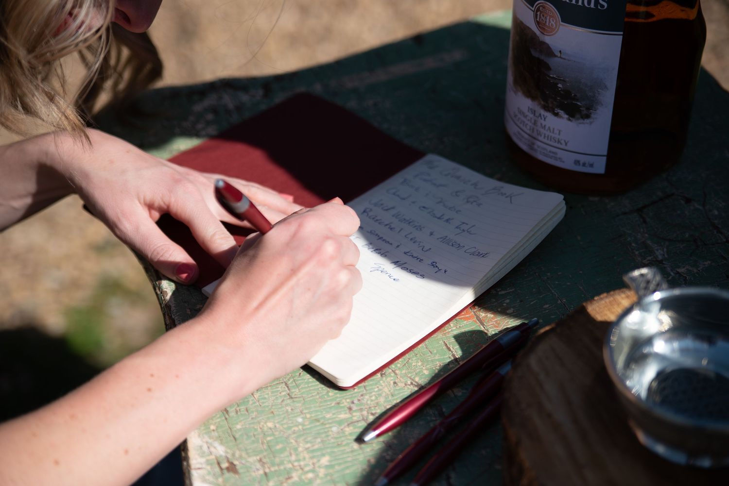 Guest signing wedding guestbook with red pen at outdoor table in warm sunlight.