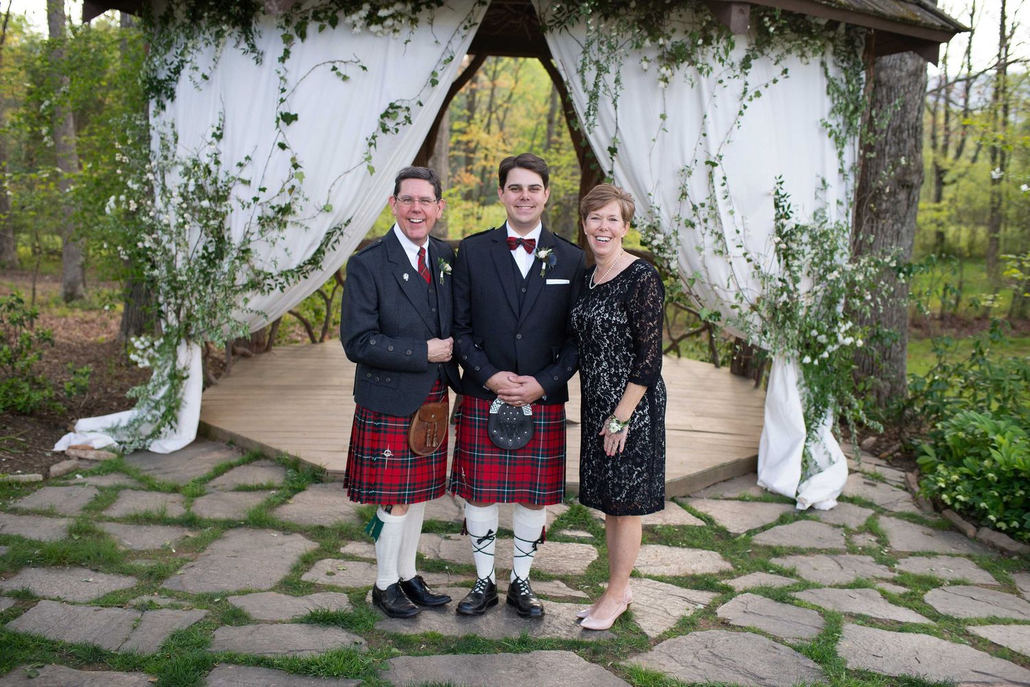 Family of three posing at outdoor wedding ceremony under floral arch, man wearing traditional Scottish tartan kilt.