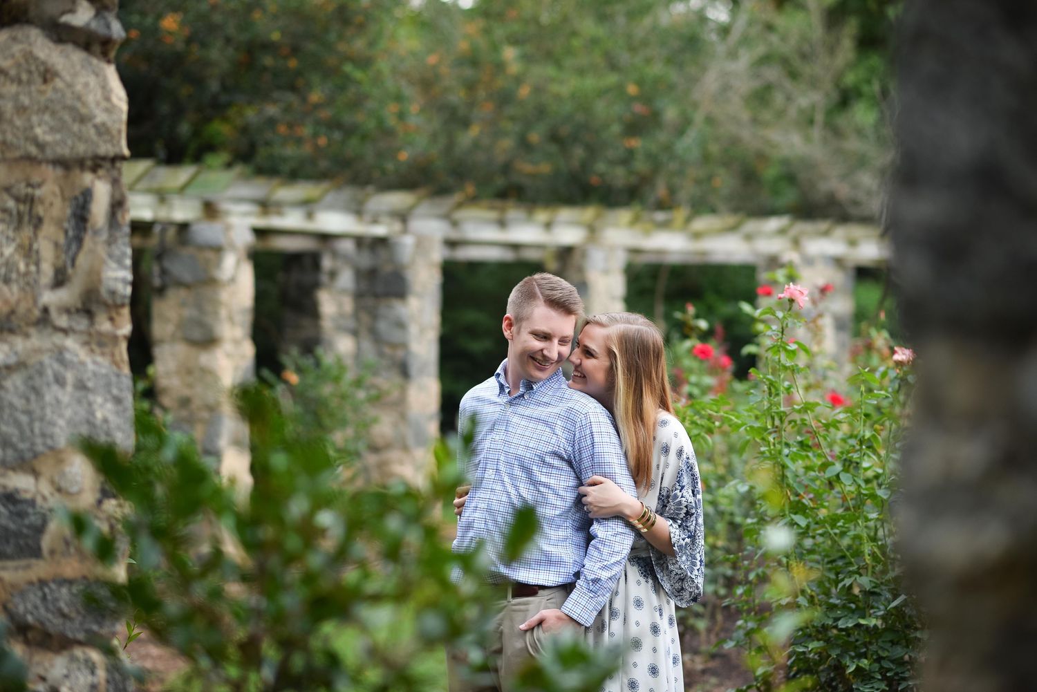 Couple embracing in a lush garden with stone columns and blooming roses in the background.