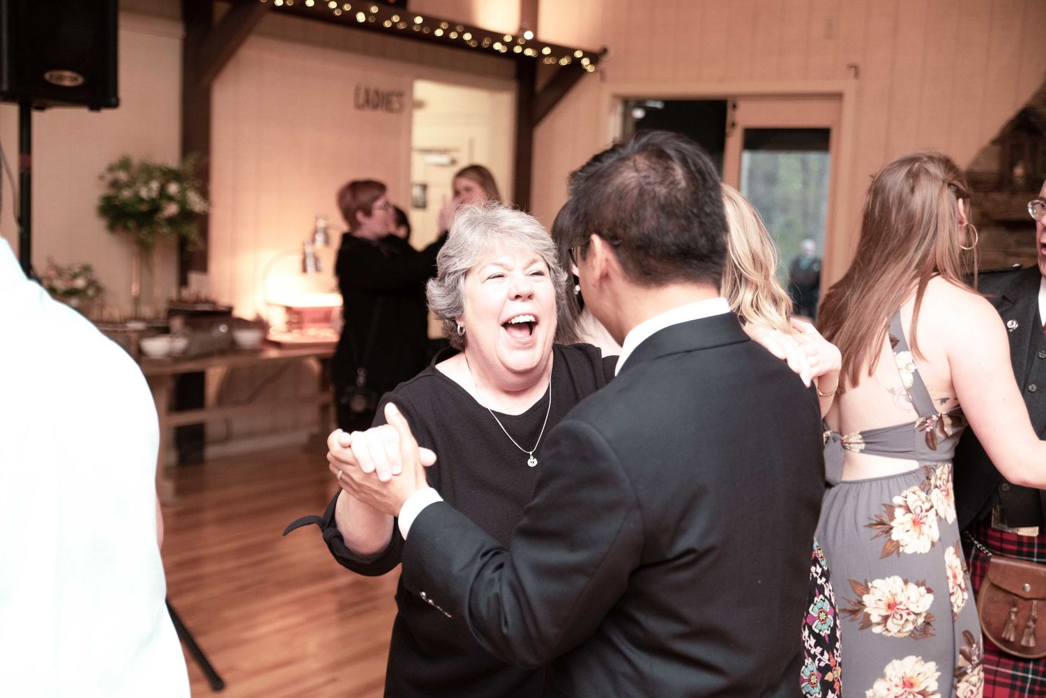 Elderly woman laughing joyfully while dancing with a man in a suit at a wedding reception in a rustic barn venue.