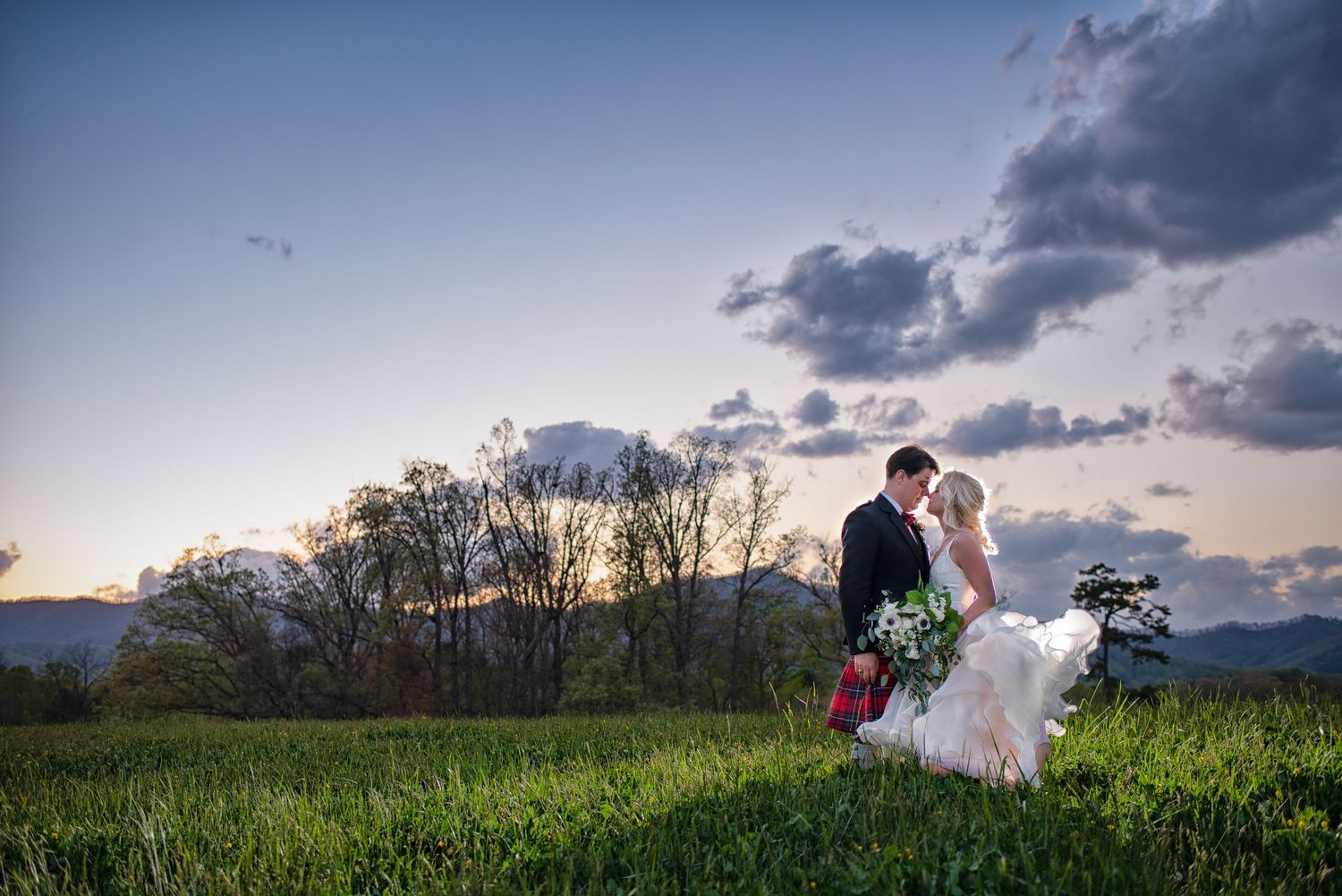 Romantic bride and groom kissing in green meadow at sunset with mountain backdrop and dramatic sky.