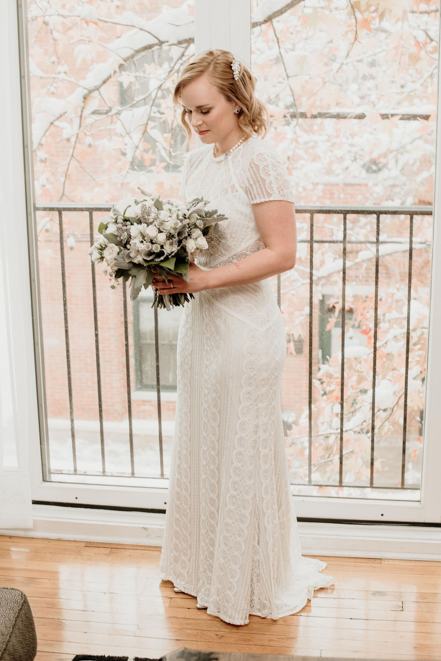 Bride in white lace gown holding floral bouquet standing by large window with snowy trees backdrop.