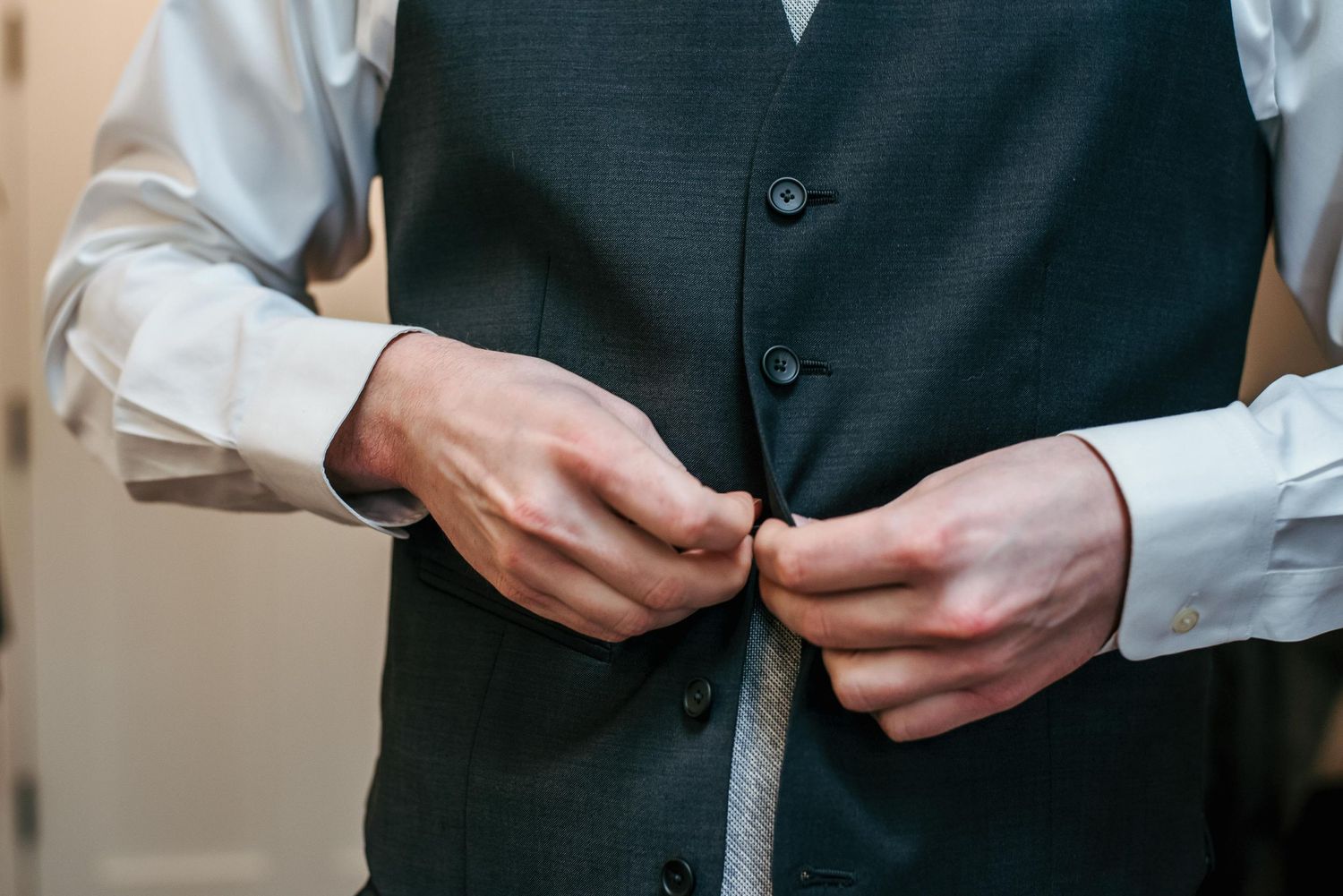 Man buttoning a dark gray suit vest over a white dress shirt, getting dressed for a formal occasion.