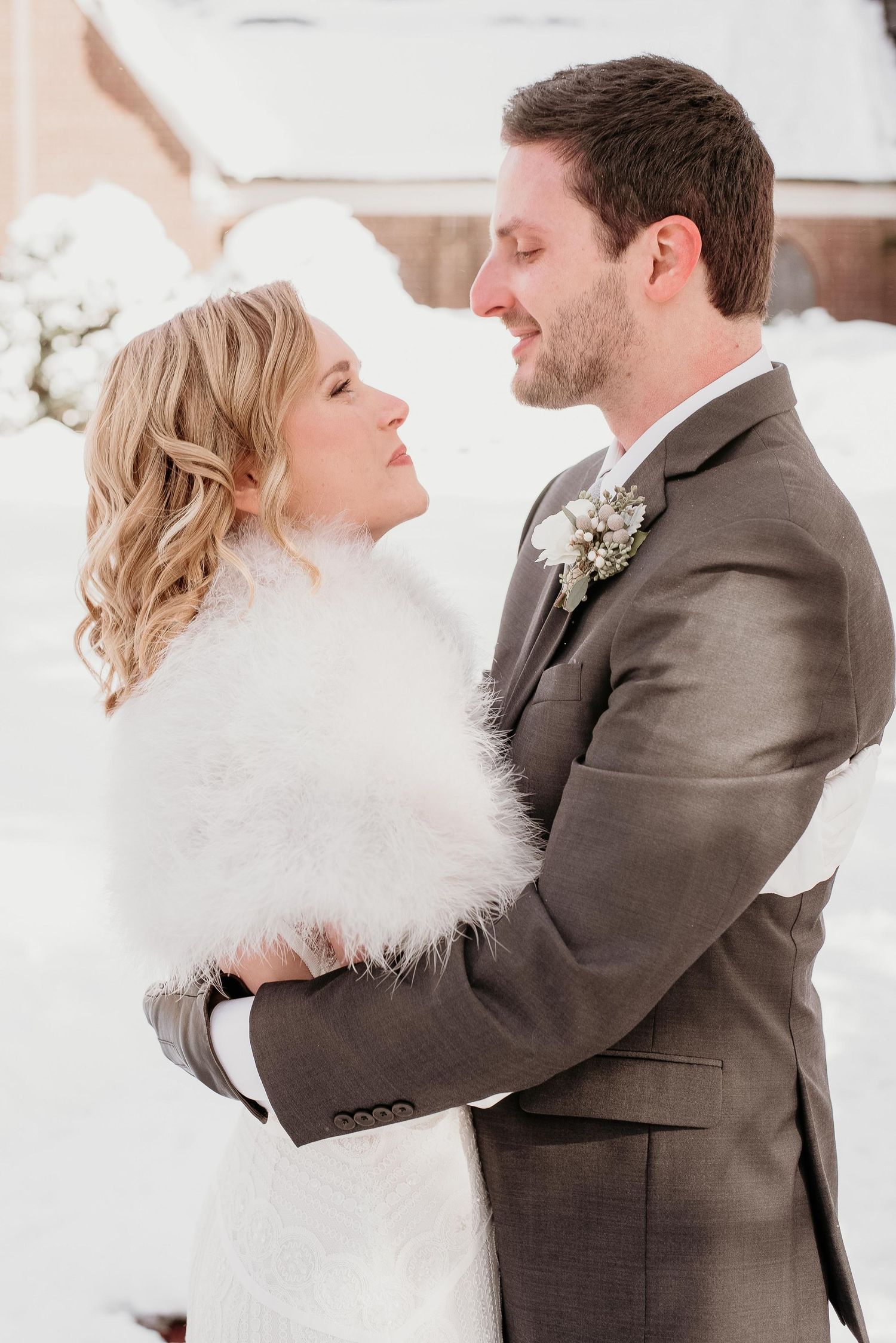 Bride in white fur wrap and groom in gray suit embrace outdoors in snowy winter wedding setting.