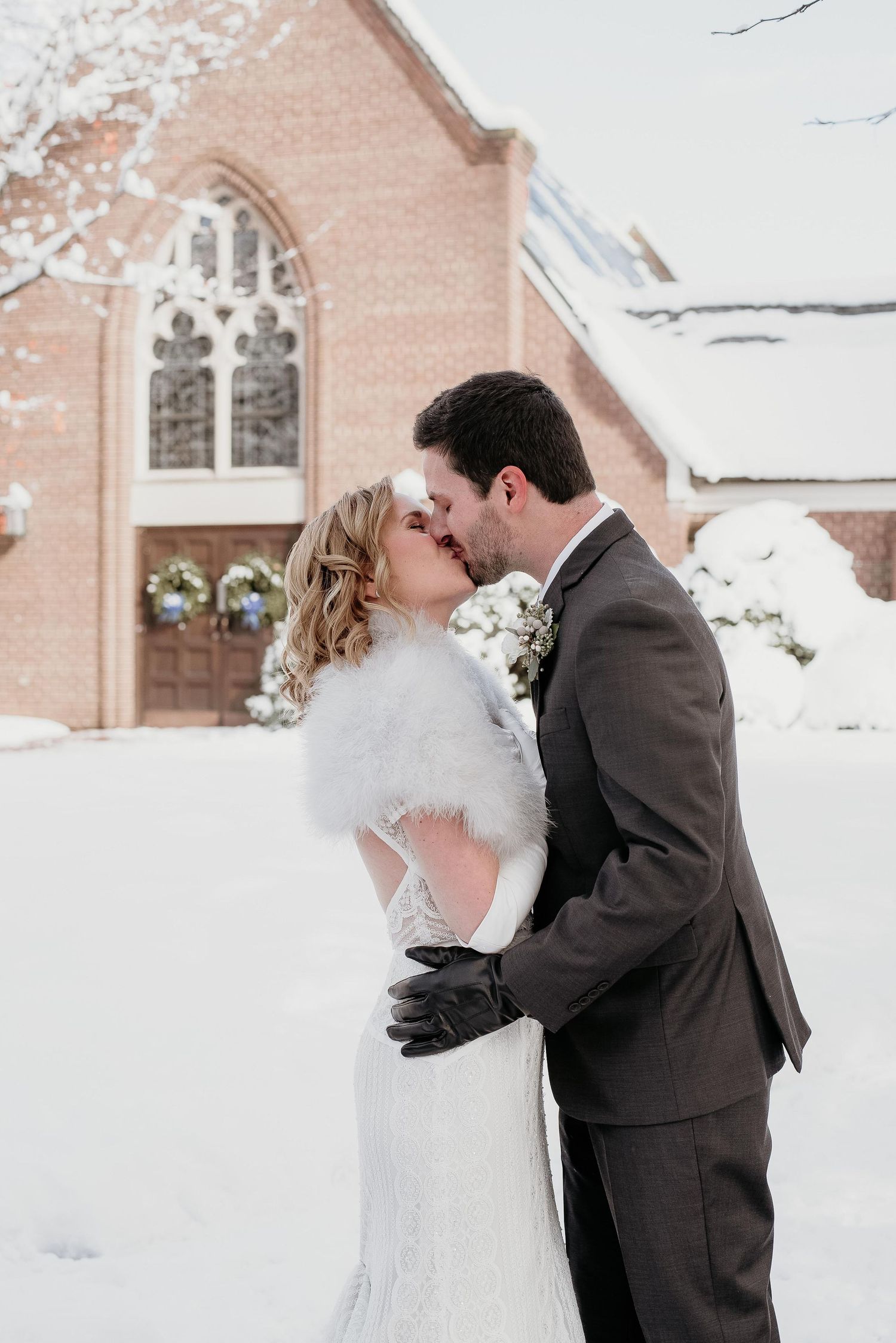 Bride and groom share a kiss in the snow outside a brick church, bride wearing a white fur stole and groom in a gray suit.