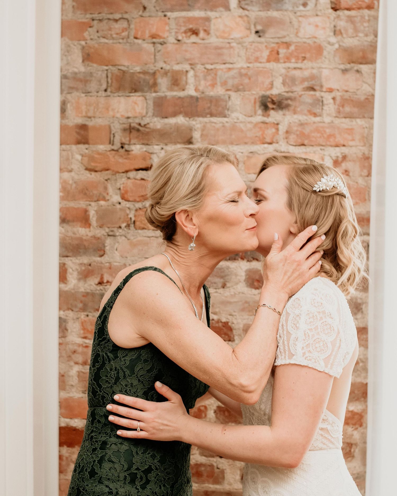 A bride in white lace receives a tender kiss from an older woman in green lace near a rustic brick wall.
