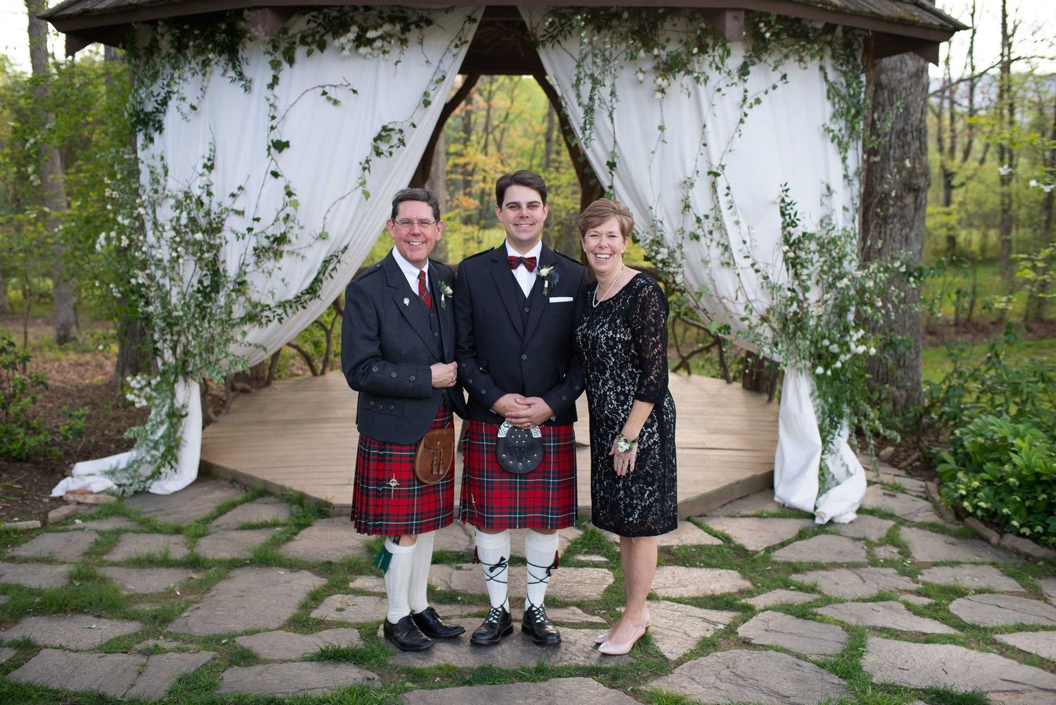 Groom in red tartan kilt poses with family under floral gazebo at outdoor wedding ceremony.