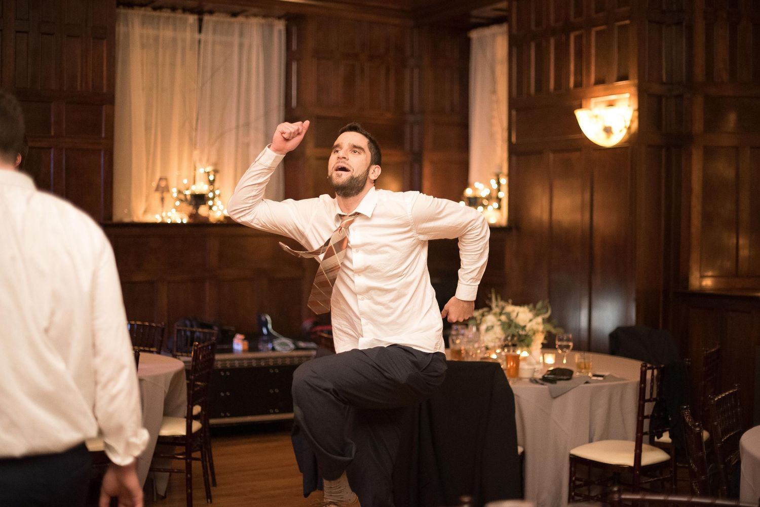 Man dancing energetically at a wedding reception in an elegant wood-paneled ballroom, wearing a white dress shirt.