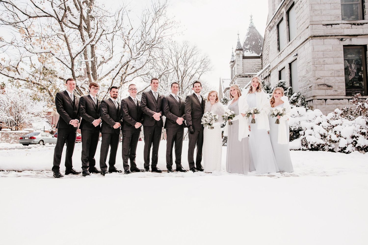 Wedding party poses in snow outside historic building, groomsmen in dark suits and bridesmaids in silver gowns.