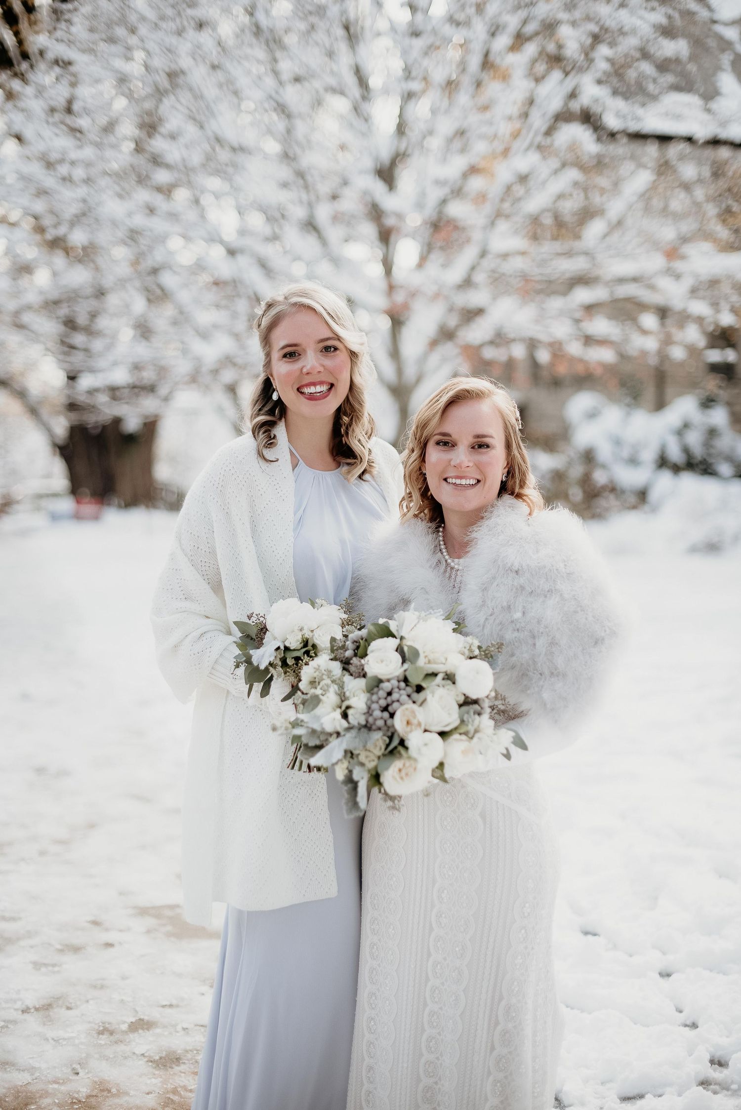 Two brides in white winter attire hold floral bouquets while posing in a snowy outdoor setting.