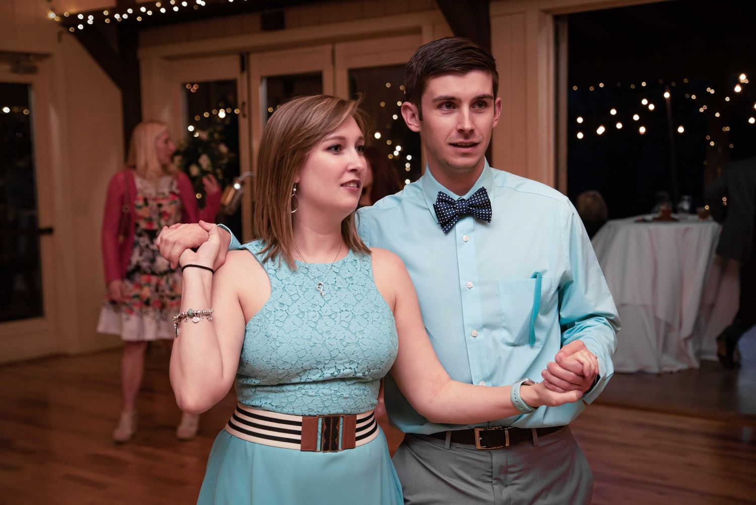 Couple in matching teal outfits dancing at a wedding reception with string lights in the background.
