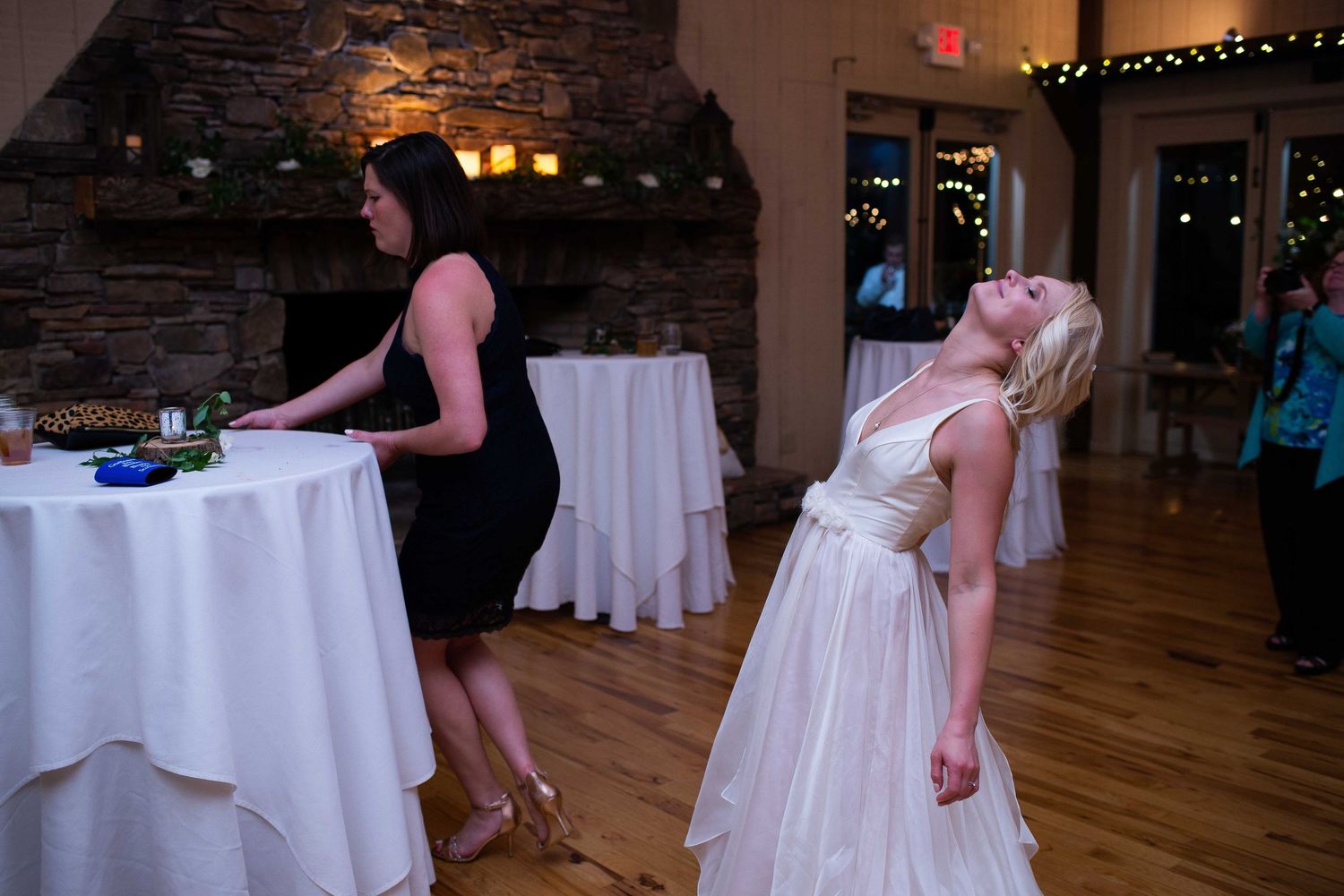 Blonde bride in white gown tilts head back at wedding reception while woman in black dress stands nearby.