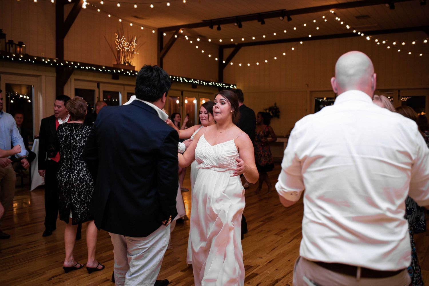 Bride dancing with guests at a rustic wedding reception venue with string lights overhead.