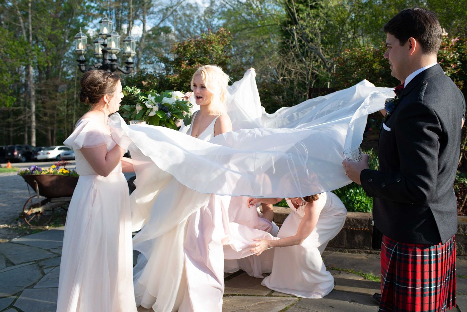 Bridesmaids helping bride with flowing white wedding dress outdoors at elegant venue with man in Scottish kilt nearby.