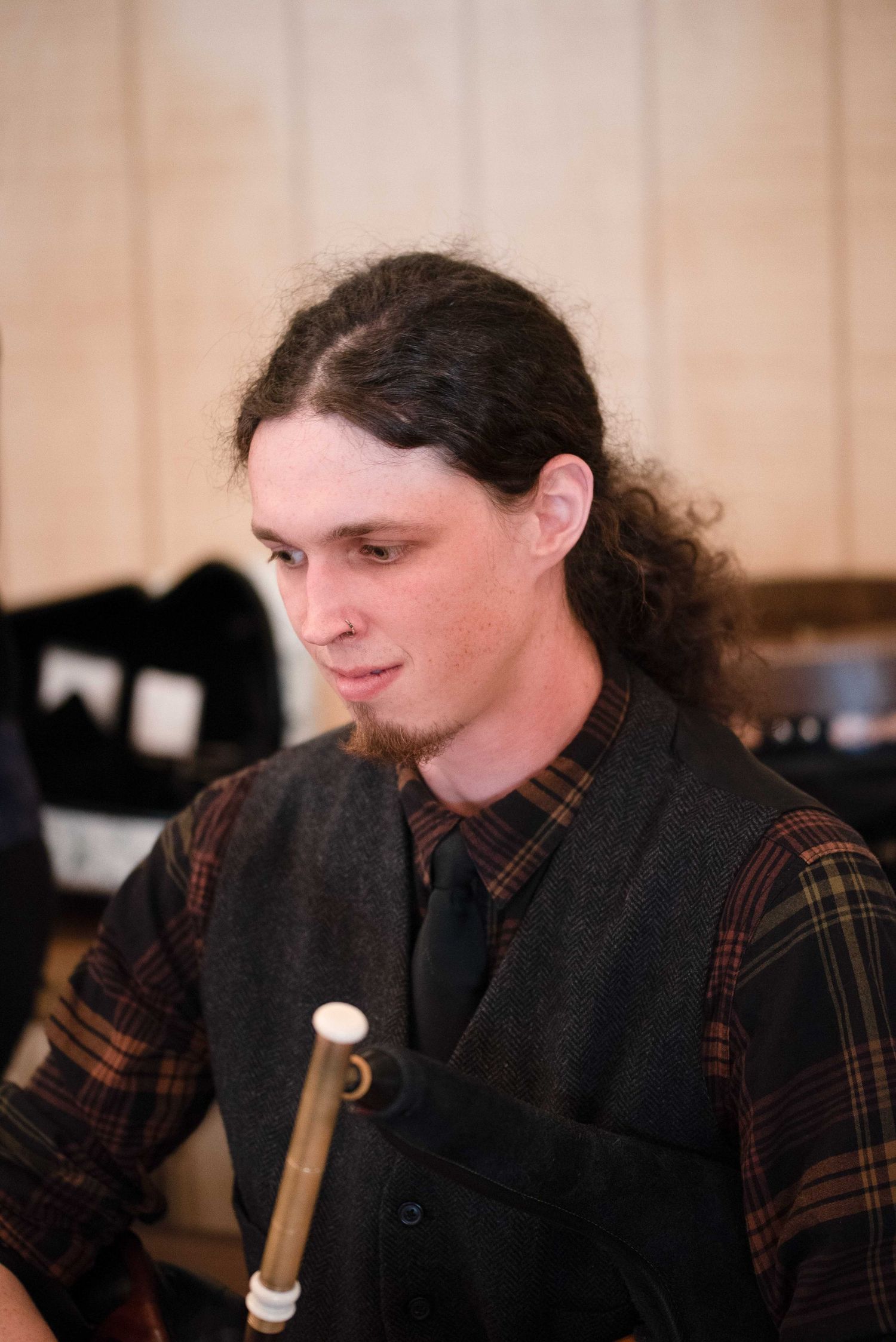 Young man with long curly hair playing bagpipes, wearing a plaid shirt and gray vest.