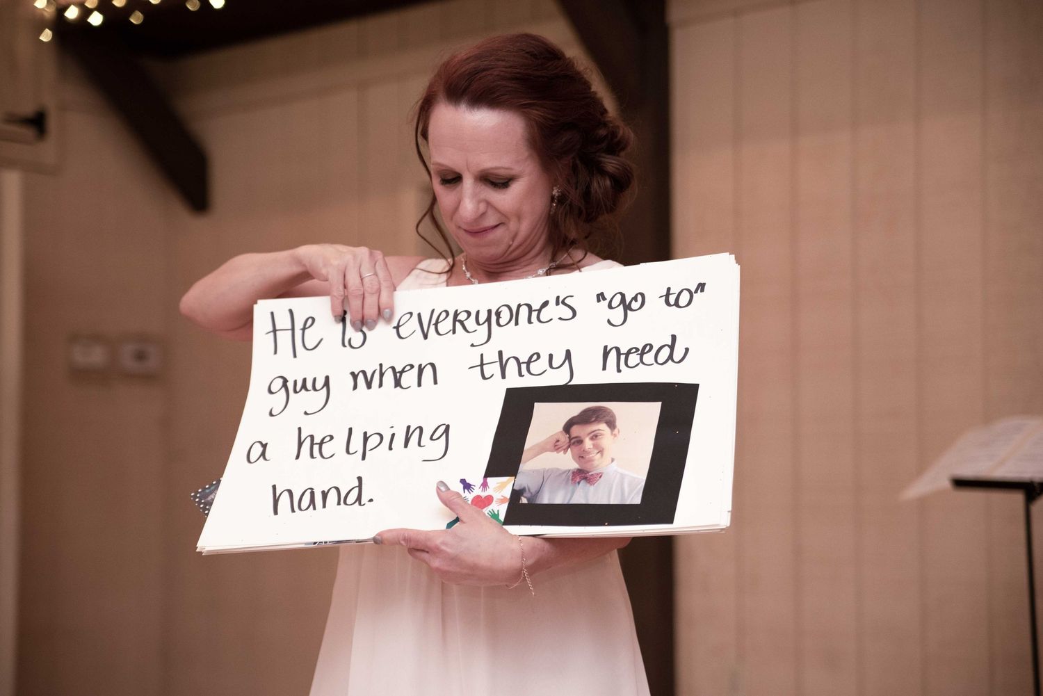 Woman holding a handwritten sign with a framed photo at a wedding reception, sign reads about someone being a helpful person.